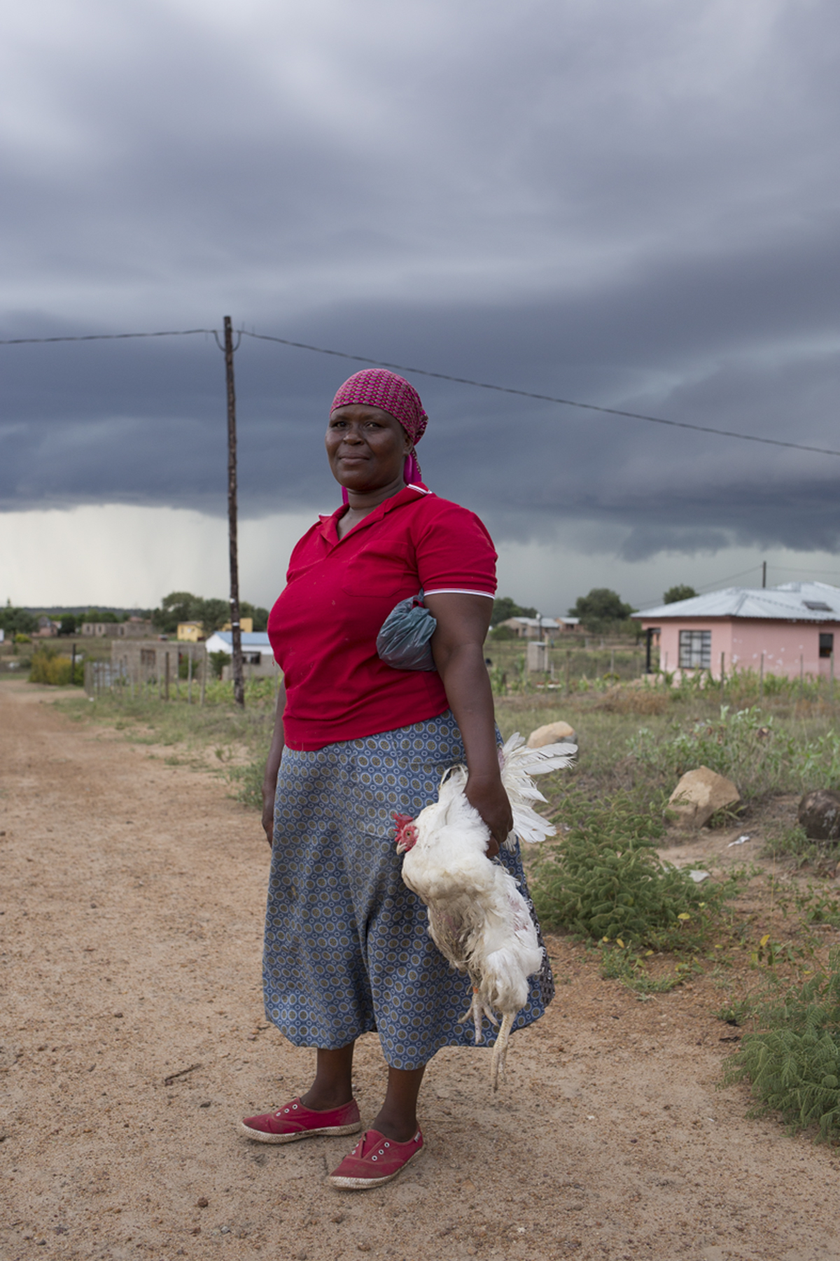 Giya Makondo-Wills, “They Came From The Water While The World Watched” – Woman with a red shirt and shoes and blue patterned skirt, holding a chicken by its wings with the left hand and walking on a dirt road in front of a row of houses and shrubs