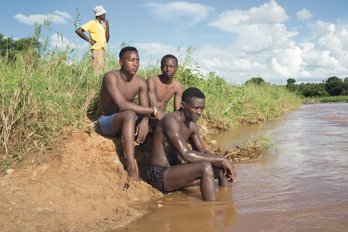 Giya Makondo-Wills, “They Came From The Water While The World Watched” – group of three young men sitting by a river in swimwear while a fourth, fully dressed man in the background looks into the distance