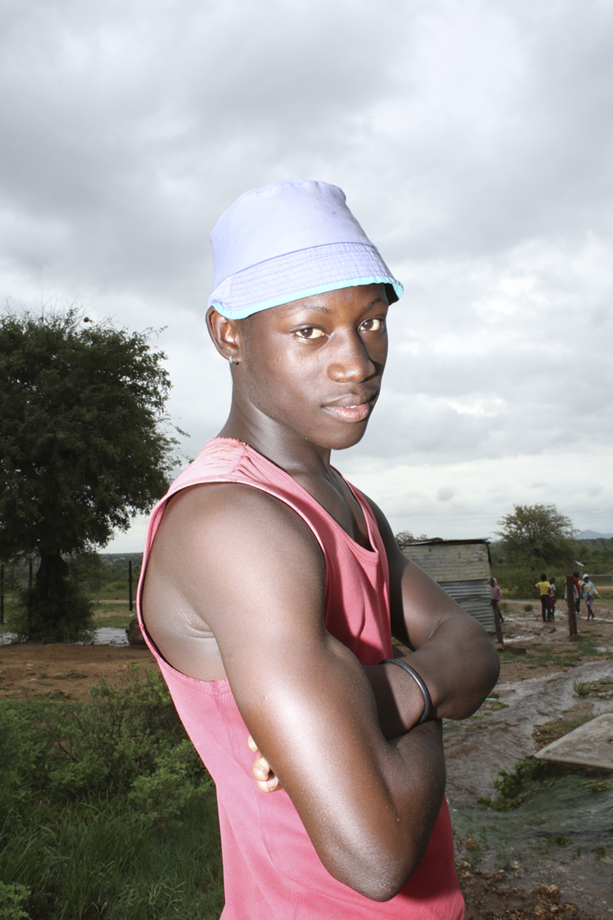 Giya Makondo-Wills, “They Came From The Water While The World Watched” – young muscular man wearing a pink tanktop and a blue bucket hat and looking at the camera from his profile