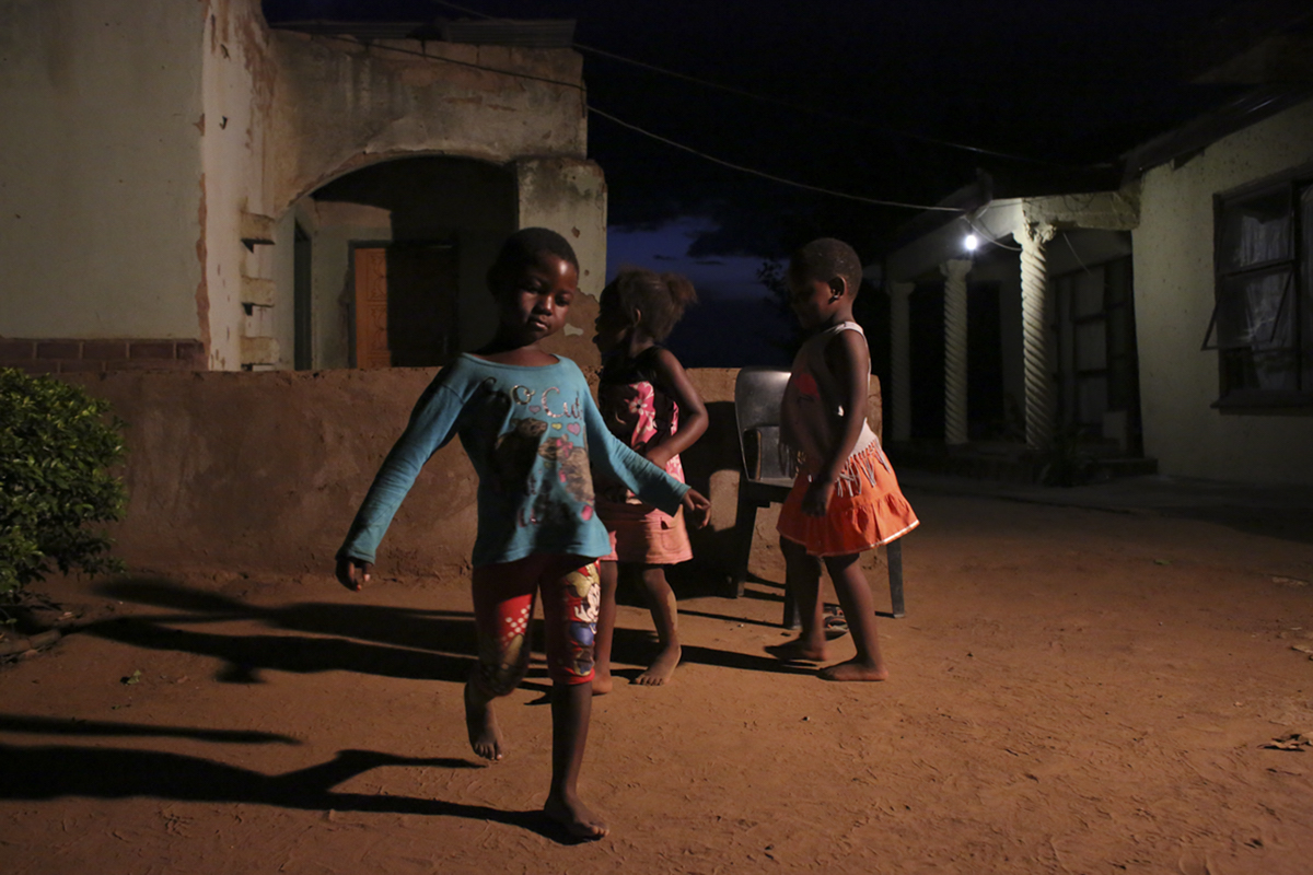 Giya Makondo-Wills, “They Came From The Water While The World Watched” – group of three children playing outside of a row of houses at night