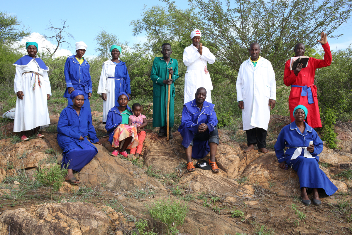 Giya Makondo-Wills, “They Came From The Water While The World Watched” – group of twelve people wearing blue, blue and white, white, red and green robes, sitting on a rock. The man in read is reading from the scriptures