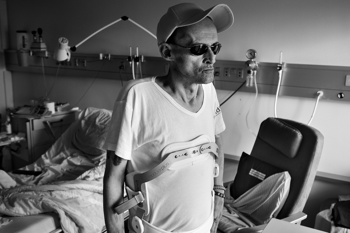 Ein abgemagerter Mann mit Sonnenbrille und Cap steht in einem Krankenhauszimmer, am Körper trägt er eine medizinische Vorrichtung