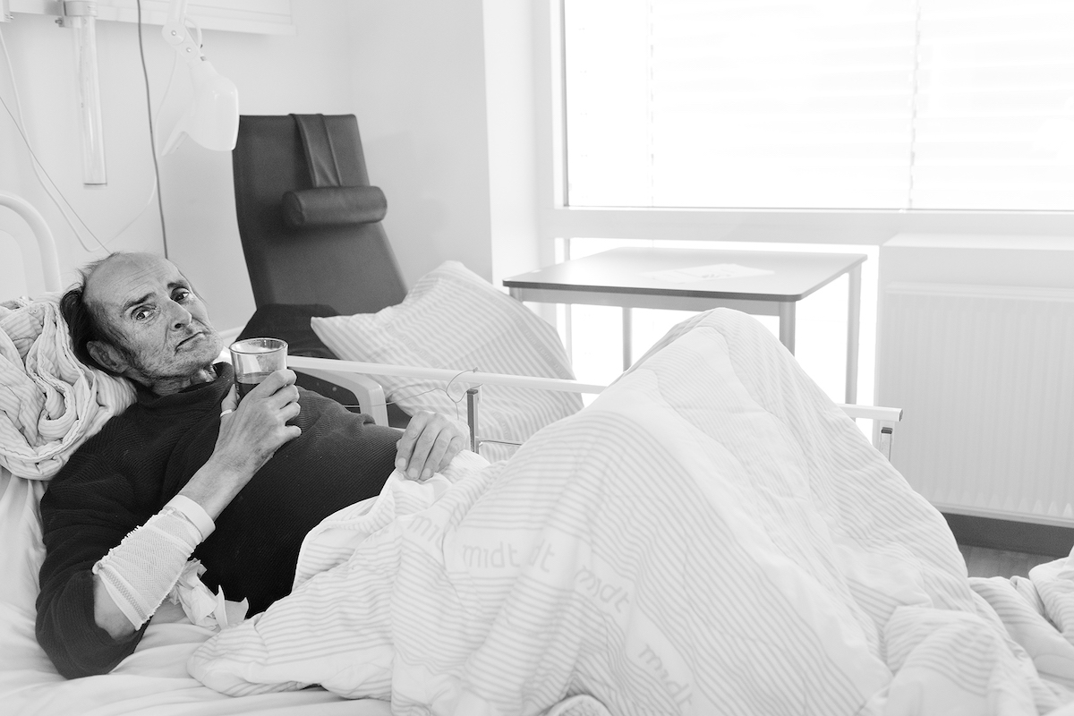 Ein alter Mann liegt in einem Krankenbett und hält ein Glas und schaut dabei in die Kamera