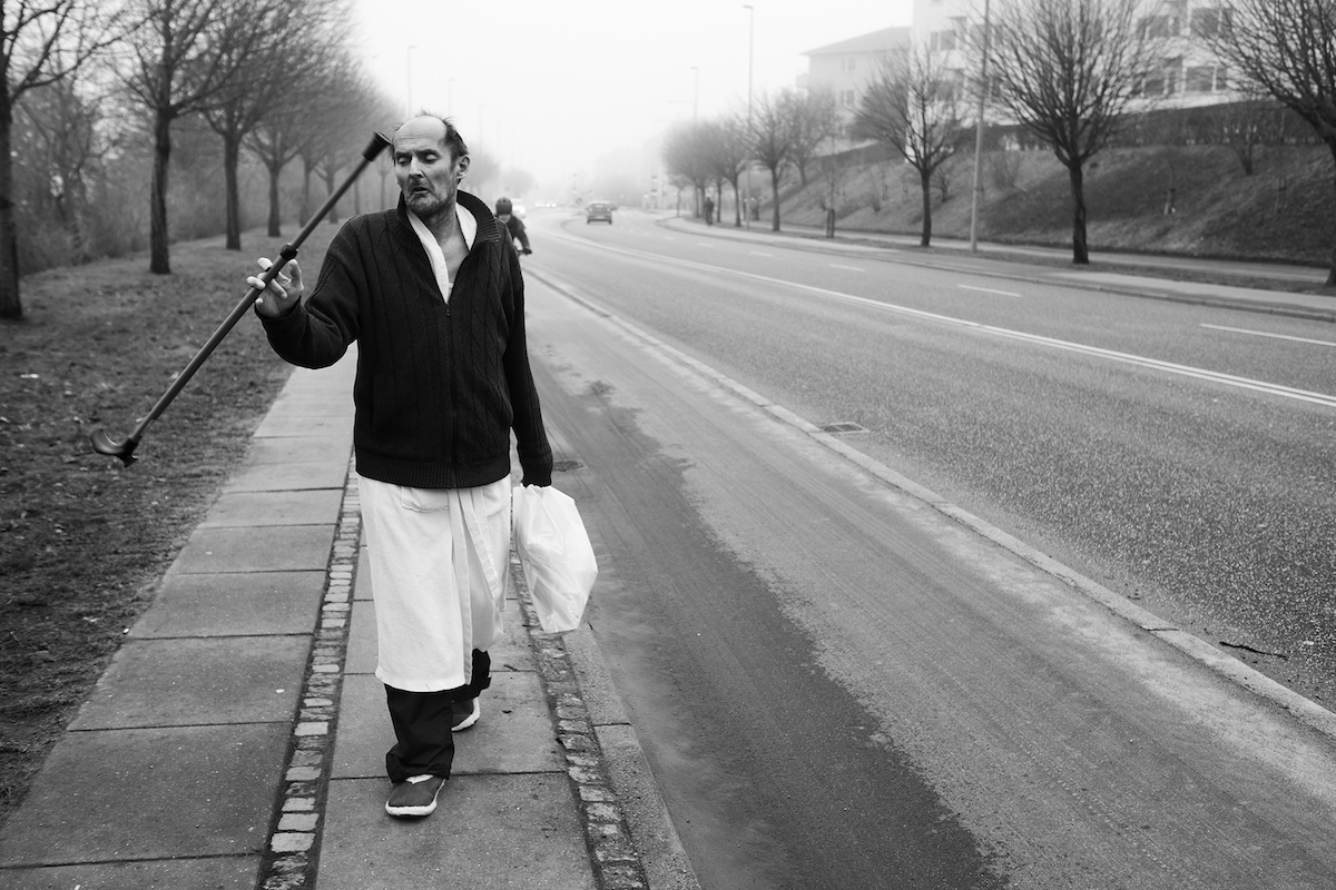 Ein alter Mann läuft in Krankenhauskleidung eine Straße entlang, in der einen Hand hält er einen Gehstock, in der anderen eine Plastiktüte