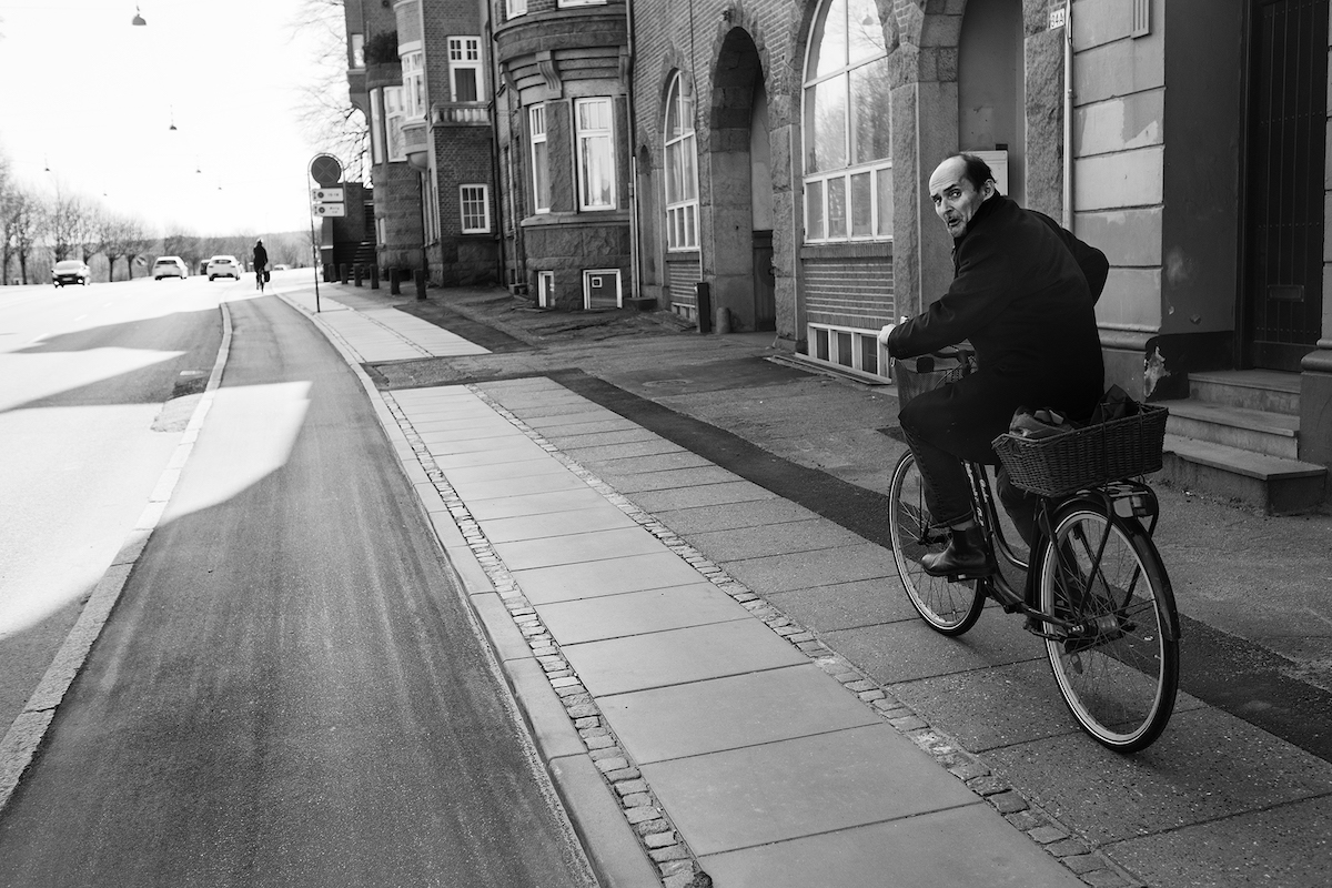 Ein Mann mit Halbglatze fährt auf dem Bürgersteig Fahrrad und schaut besorgt über seine Schulter in die Kamera