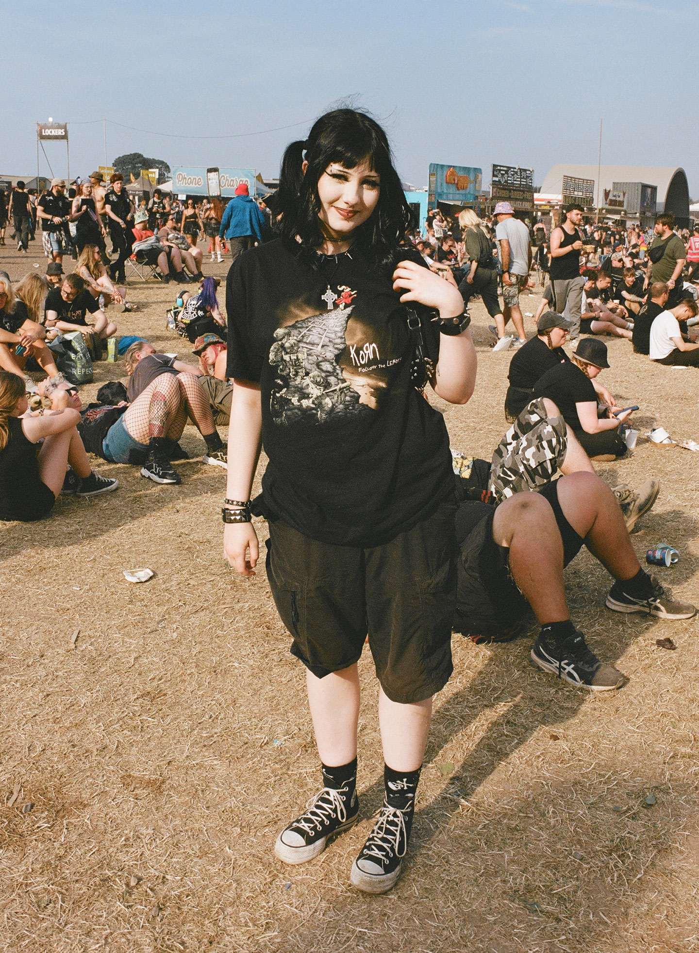 a teen girl with black hair in pigtails, a korn T-shirt and baggy shorts with converse stands in a busy festival field 