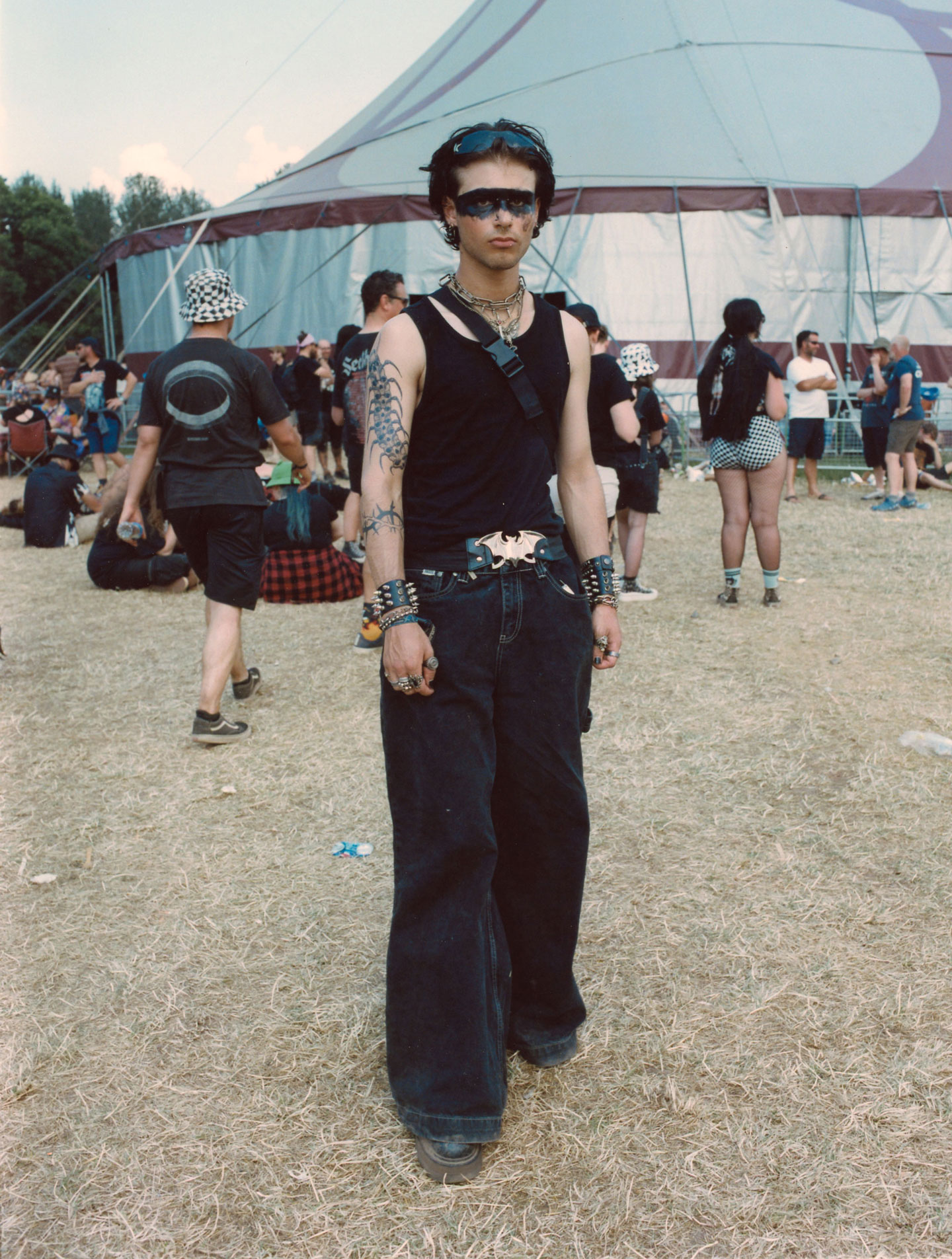 a teen boy with tattooed arms, baggy black jeans, a black tank top and a mask-like stripe painted over his eyes stands in front of a festival tent