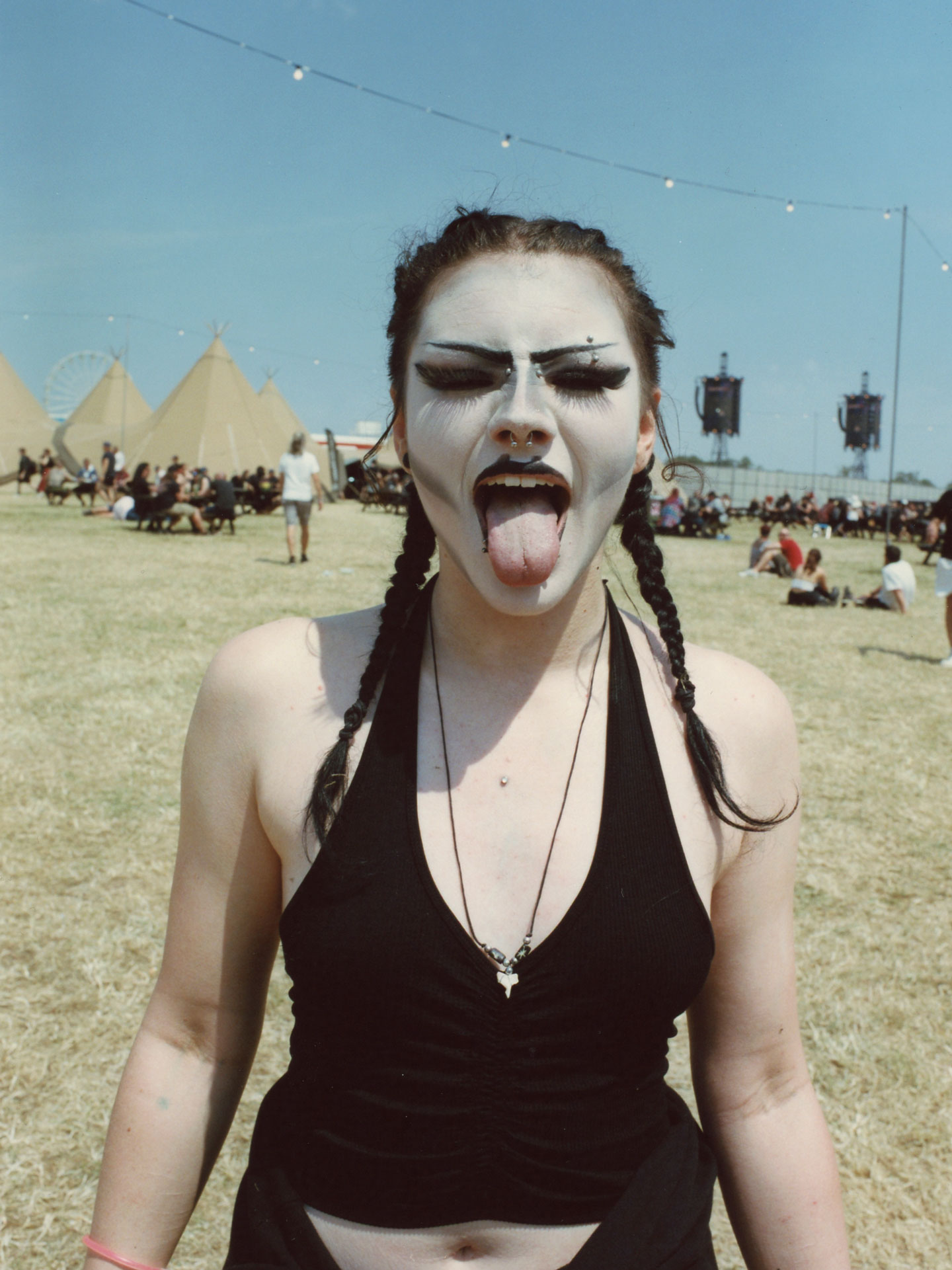 a teen girl with plaits and white gothic face paint sticks her tongue out to camera; she is standing in a festival field with tipis and dried grass behind her