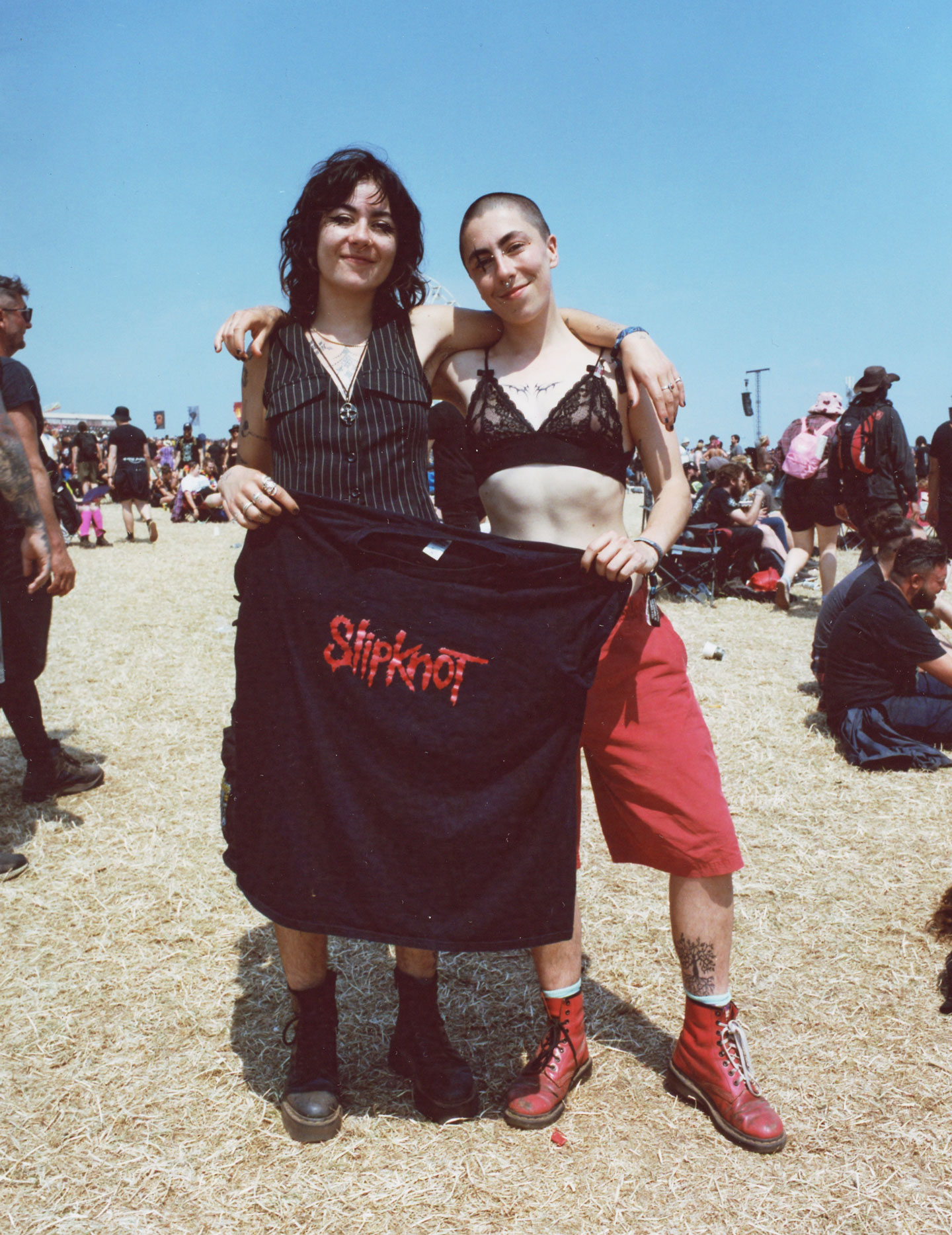 siblings wearing doc marten boots with gothic clothing (one in a pinstriped waistcoat, the other in a lace bra with a shaved head) hold a Slipknot tee between them