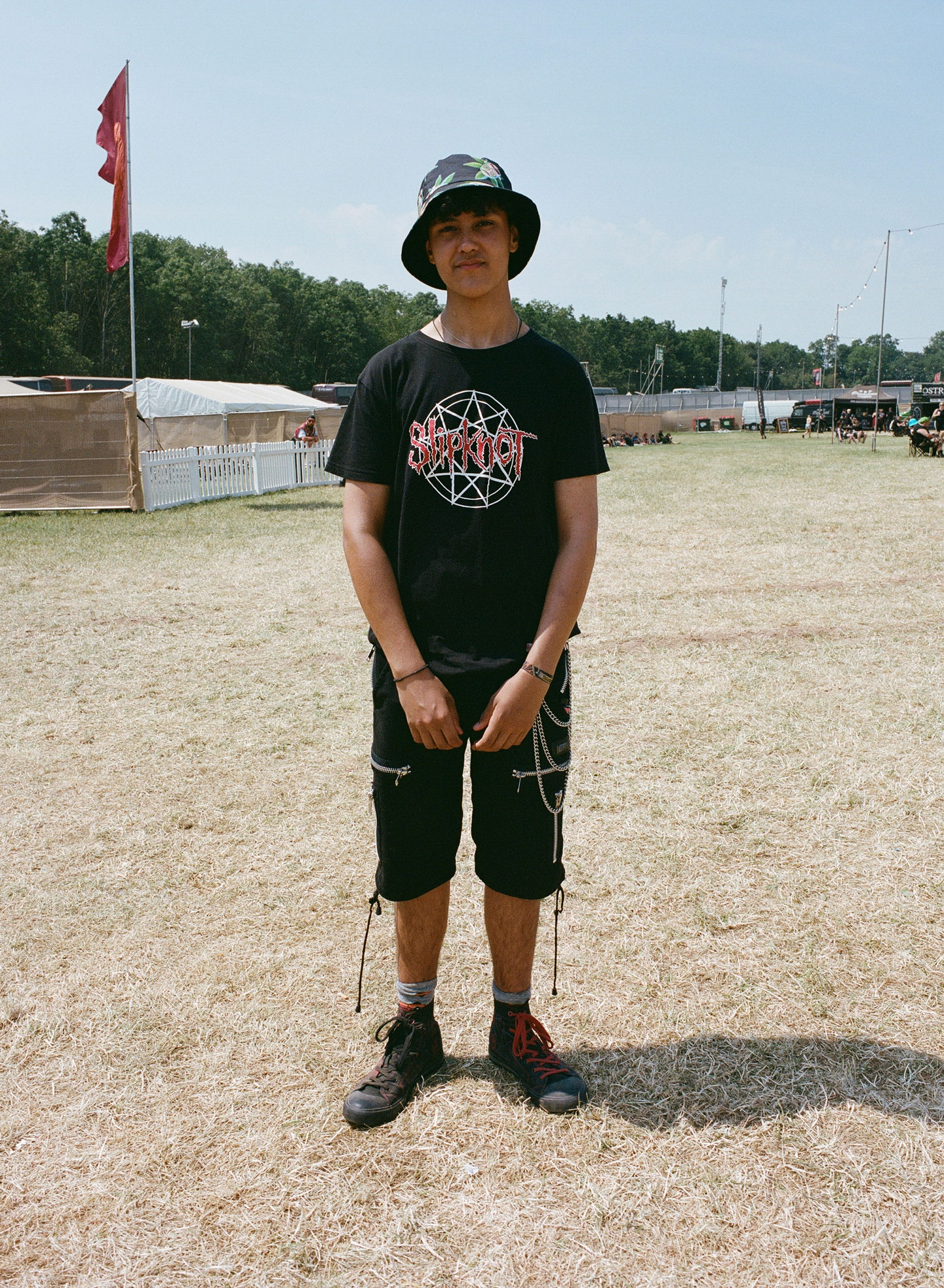 a teen boy wears baggy trousers with chains, a slipknot tee and a bucket hat stands in a dried-out festival field 