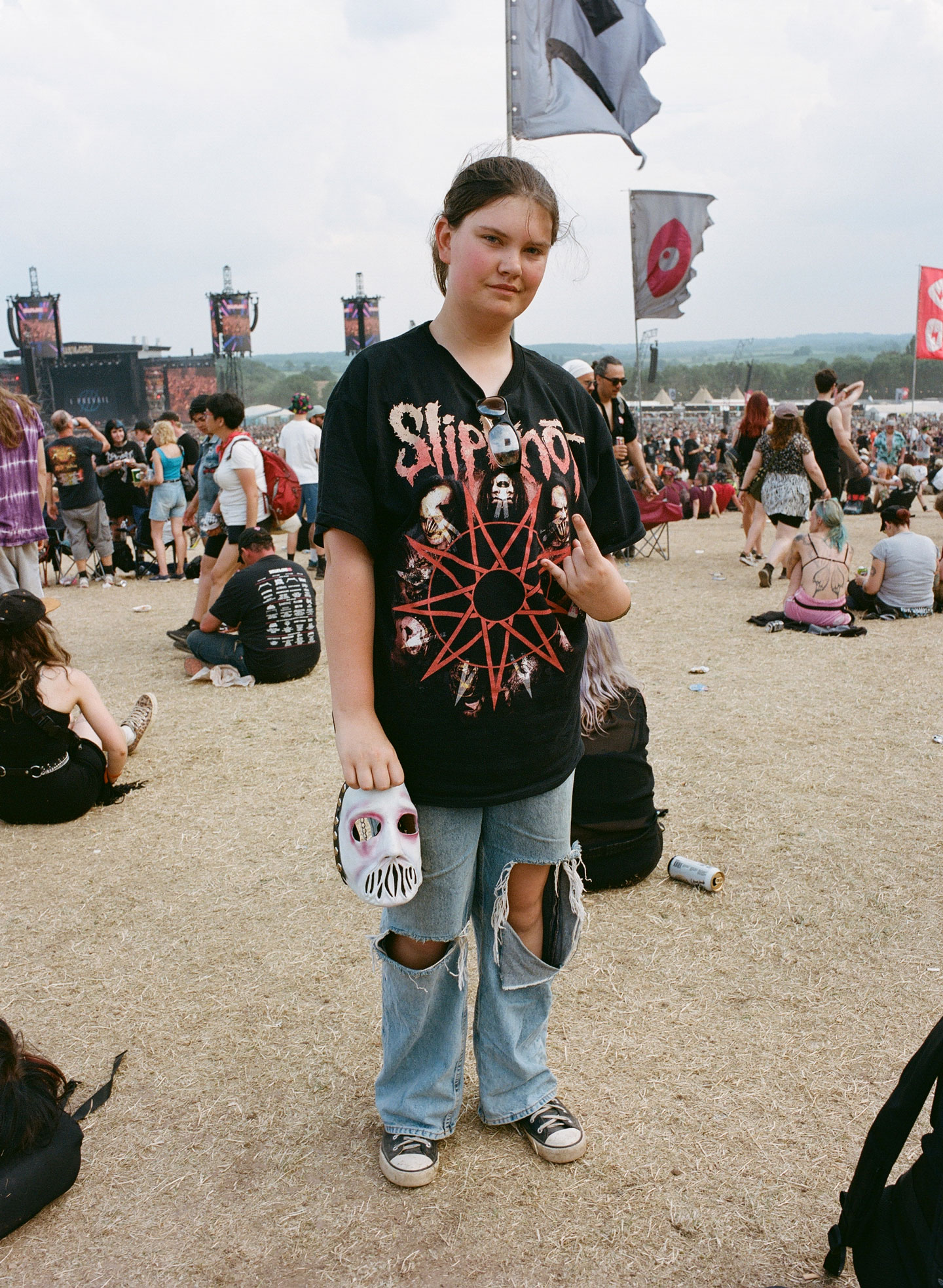 a teen brunette girl wears an oversized slipknot tee and baggy ripped jeans with converse; in her hand is a slipknot mask