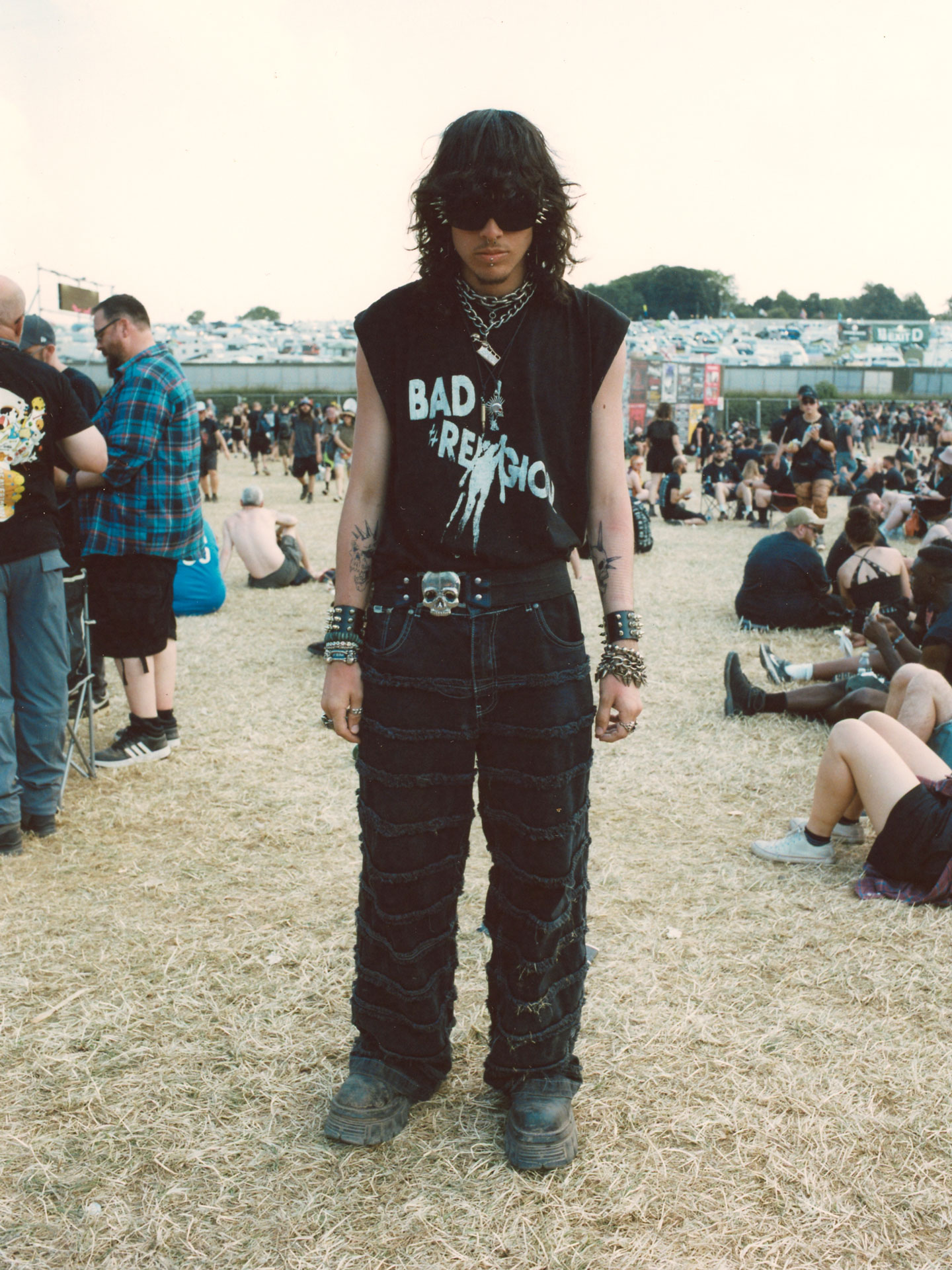 a young man with a shag haircut and a bad religion tank wears chain jewellery around his neck and wrists, plus a skill belt buckle