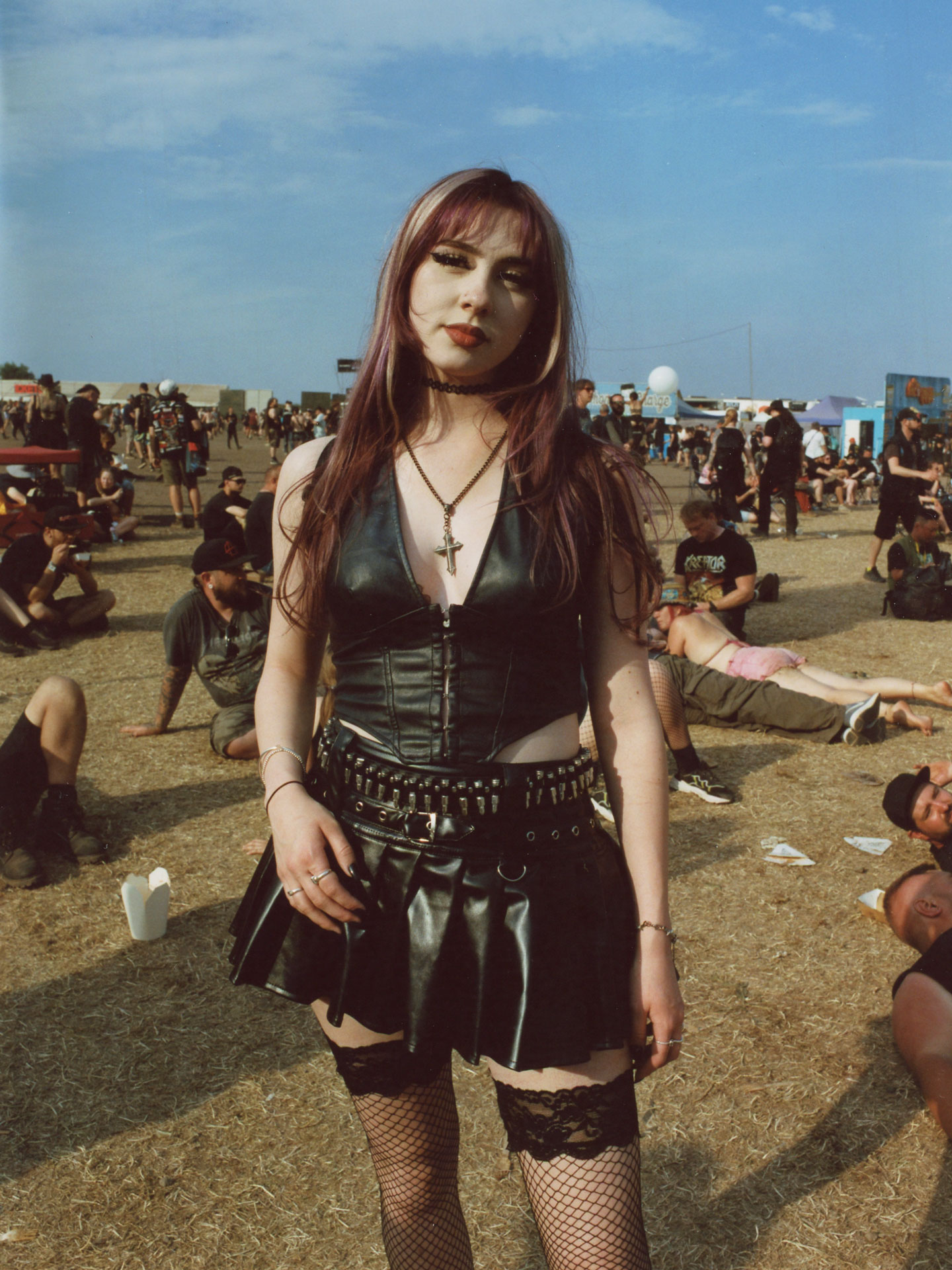 a goth girl in a leather corset and skirt wears fish net suspenders and a crucifix necklace. her hair is purple and silver 