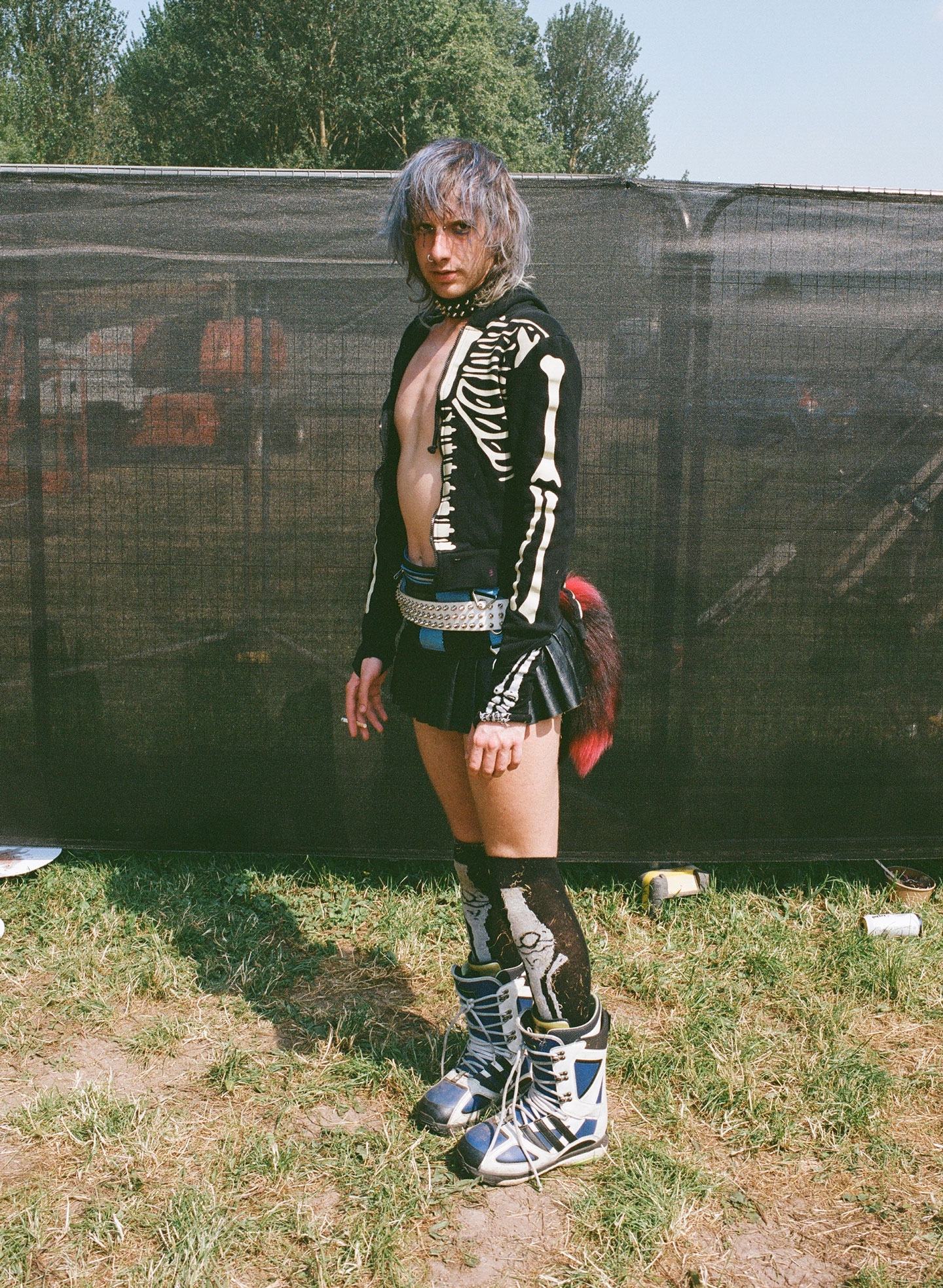 a blue-haired festival-goer wears an open skeleton tee with a short leather skirt and a red tail; they hold a cigarette in one hand