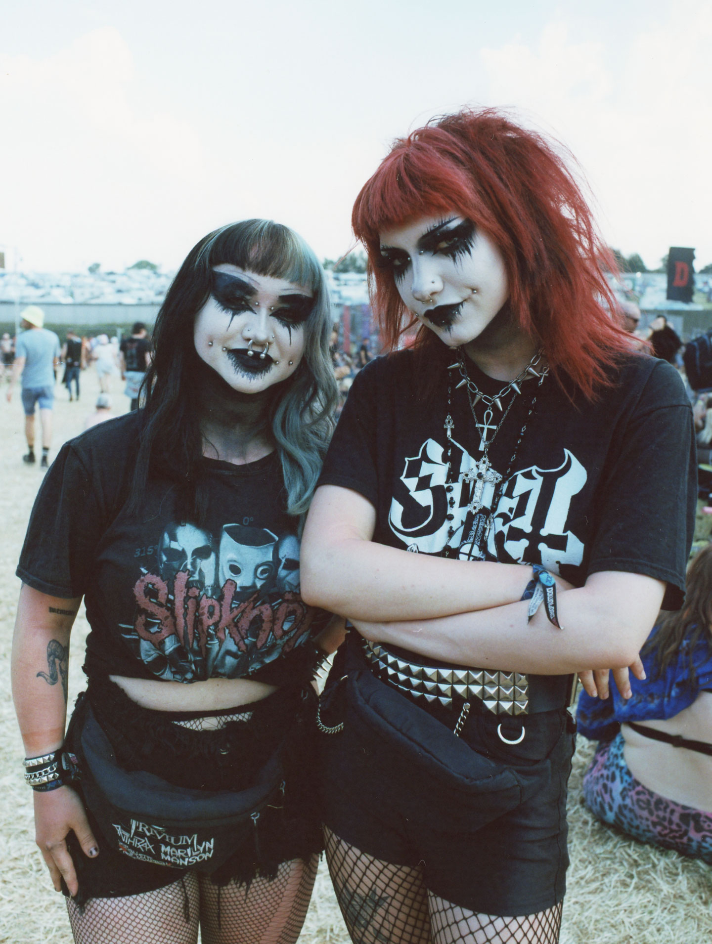 two scene kids with hair dyed red and blue stand side-by-side. their faces are painted white and they have gothic eye makeup and a lot of facial piercings