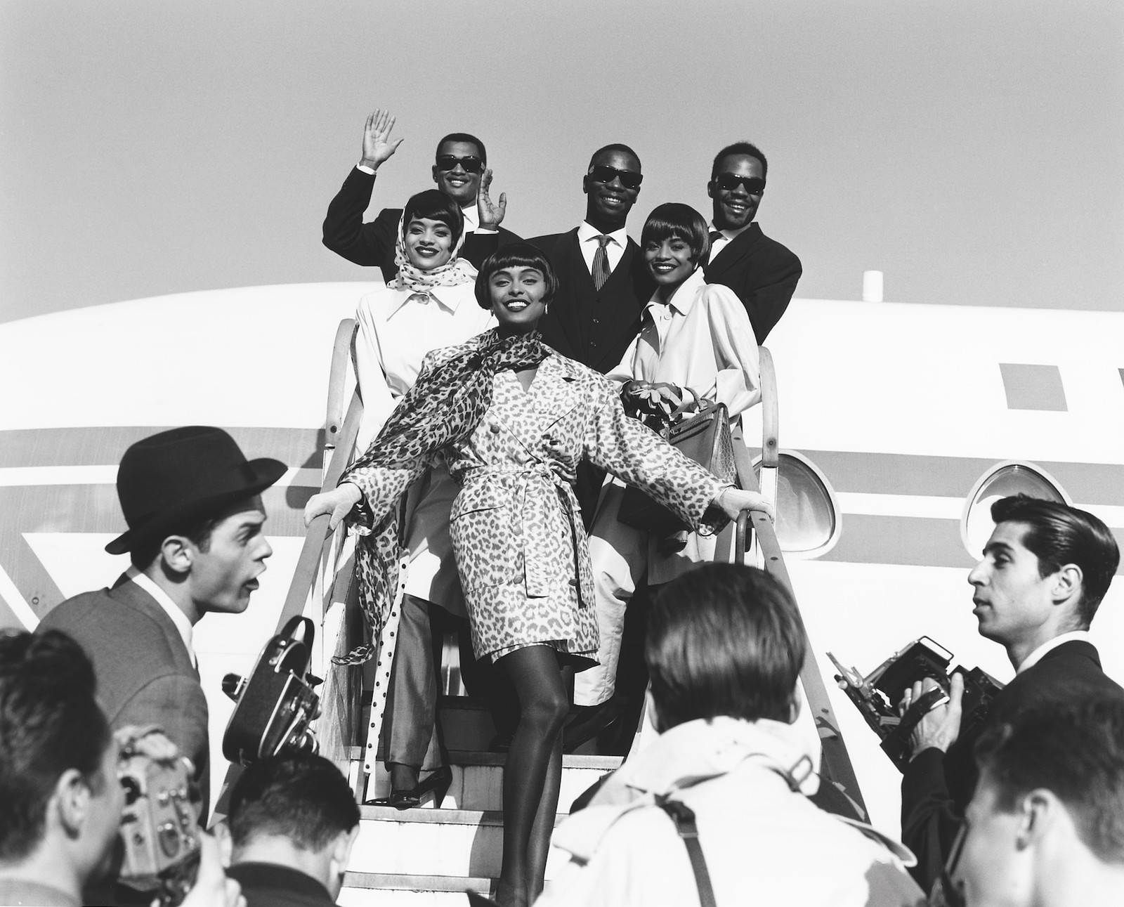 black-and-white image of models descending from an airplane as journalists wait on the steps