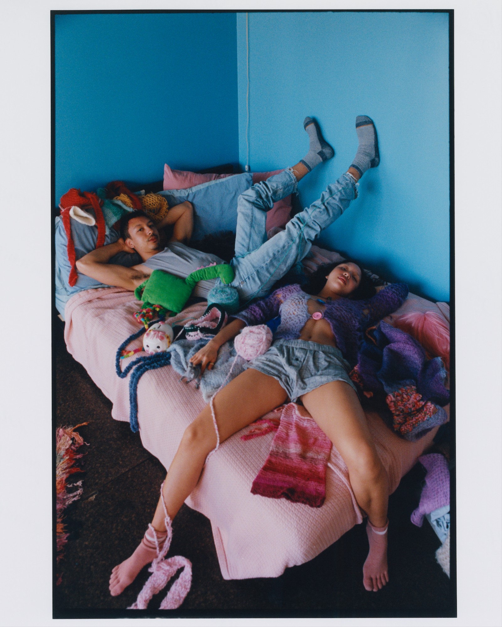 young adult twins recline on a pink double bed spread covered in colourful objects
