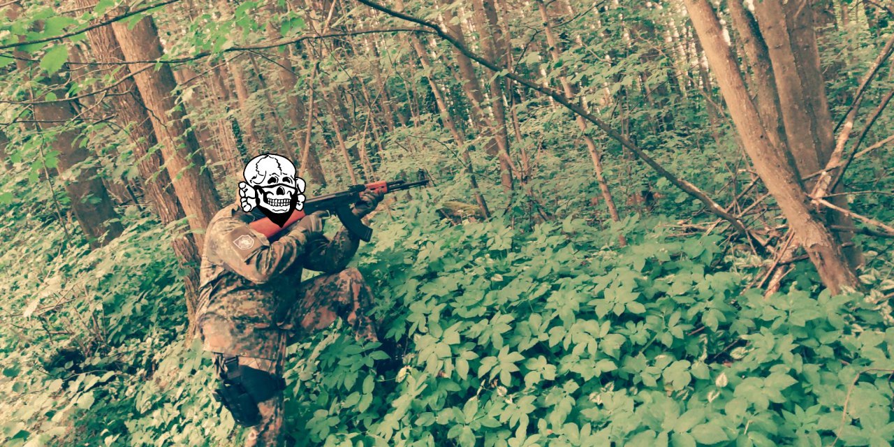 Eine Person mit Tarnanzug und gezogenem Maschinengewehr im Wald, der Kopf mit einem Totenkopf verdeckt