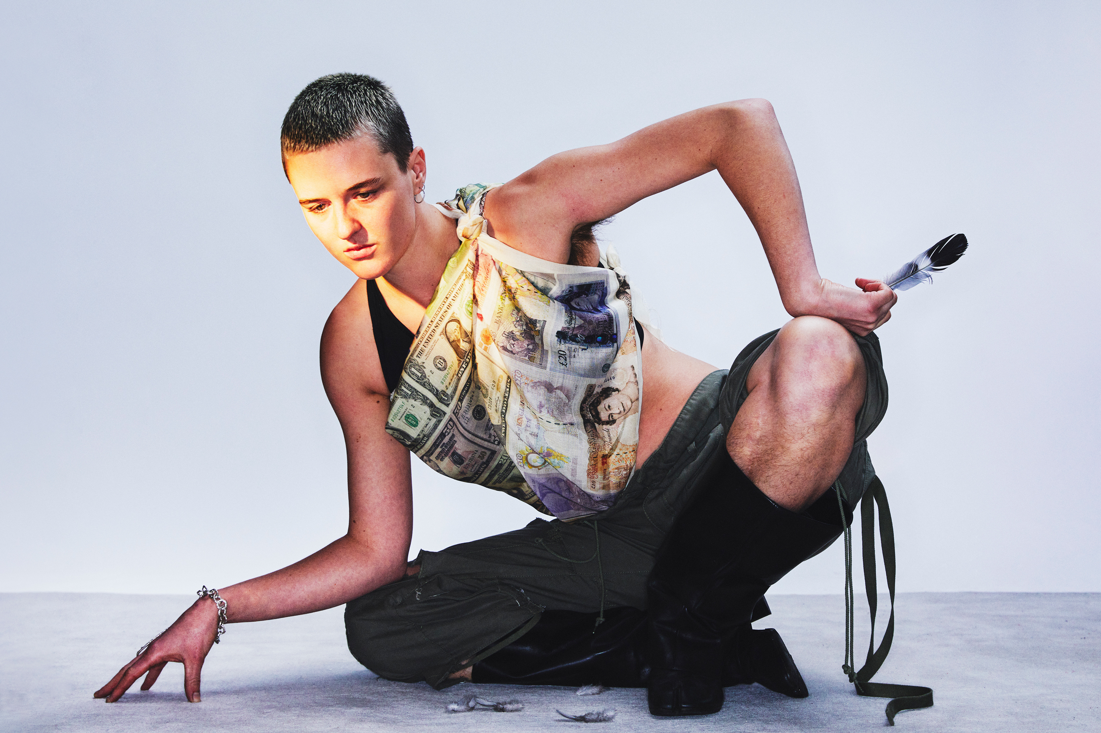 the musician tiberius b with a buzzed blonde-brown haircut, crouched on the ground in a creative pose, wearing a silk scarf printed with british pound notes as a top, shorts and black socks. they carry a pigeon feather in their hand.
