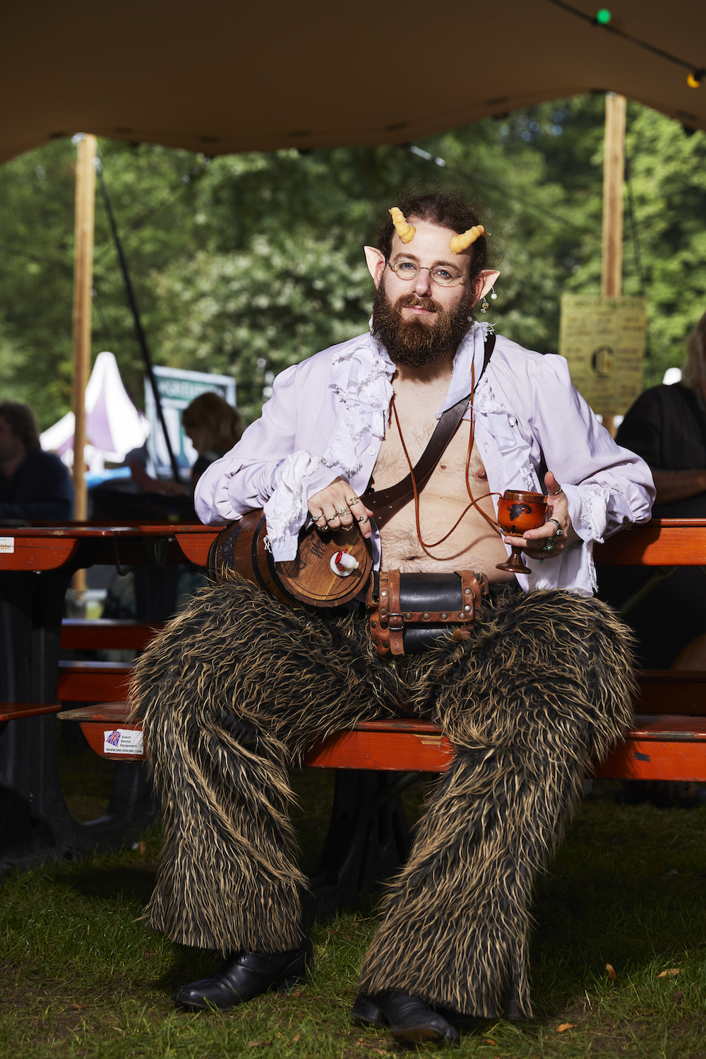 Ein Mann mit Bart, Hörnern und Fellhose sitz mit offenem Hemd auf einer Bank und hält ein Weinglas, er hat ein kleines Fass umgehangen
