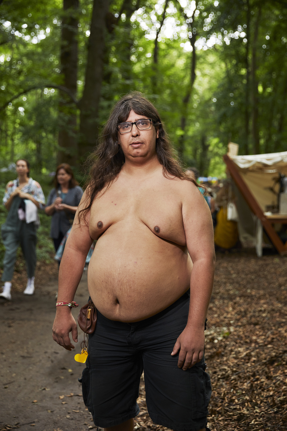 Ein übergewichtiger Mann mit langen dunklen Haare und Brille steht oberkörperfrei in einem Wald