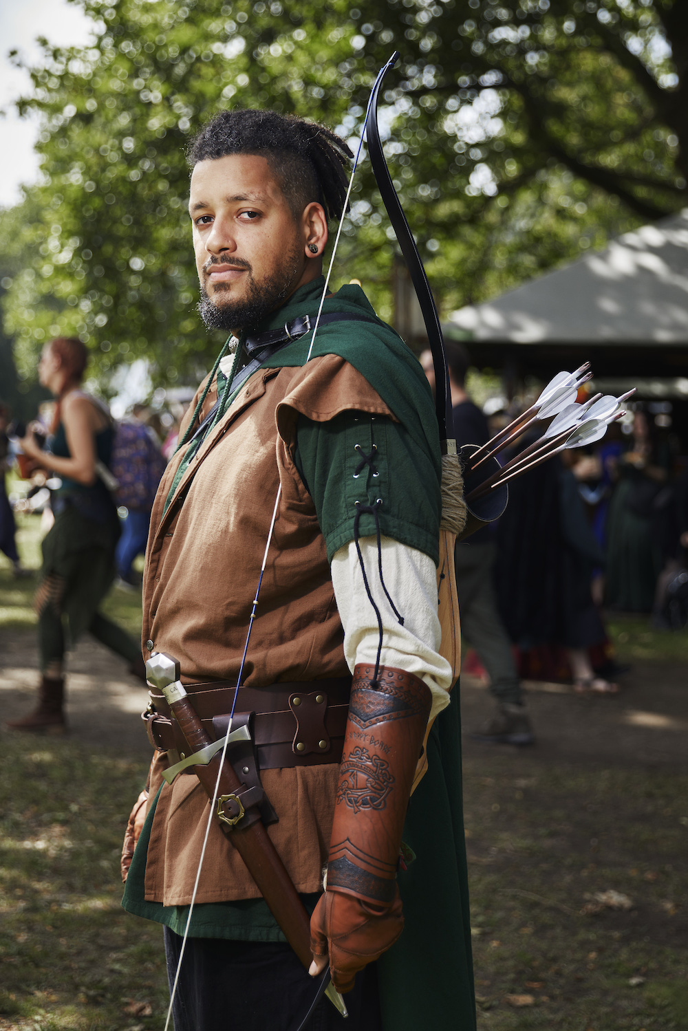 Castlefest medieval festival - man with dar skin and eyes, and dreadlocks, he's dressed in brown and green and carries a bow with arrows, a sward and leather accessories