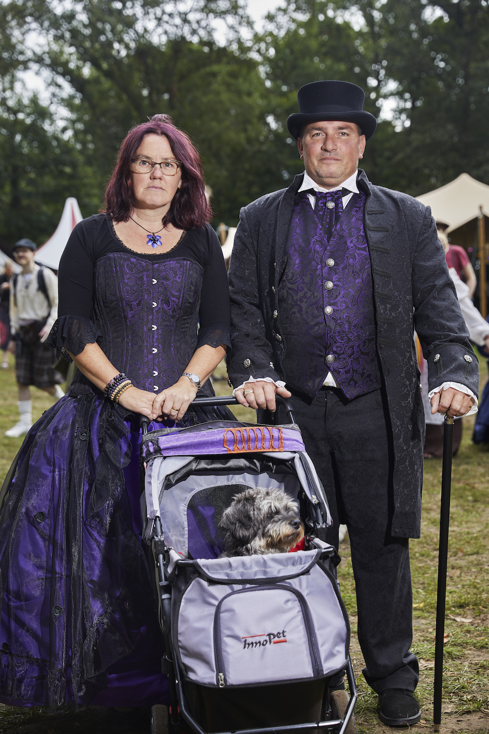 Ein Mann und eine Frau in violett-schwarzen viktorianischen Outfits vor einem lila Trolly mit einem Hund drin