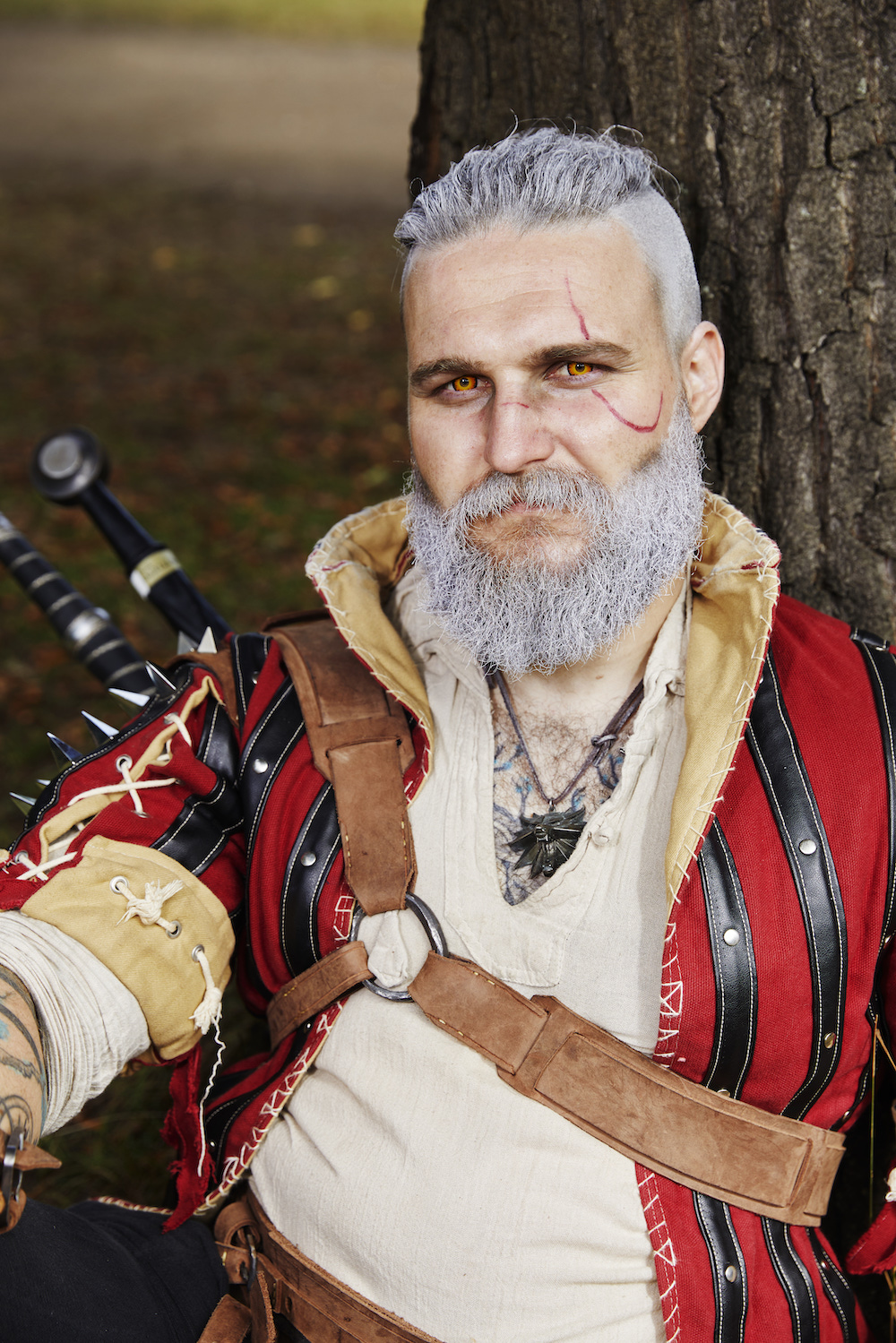 Ein Mann mit weißgefärbten Haaren und Bart und einer aufgemalten Narbe, der ein rot-schwarzes Mittelalteroutfit trägt