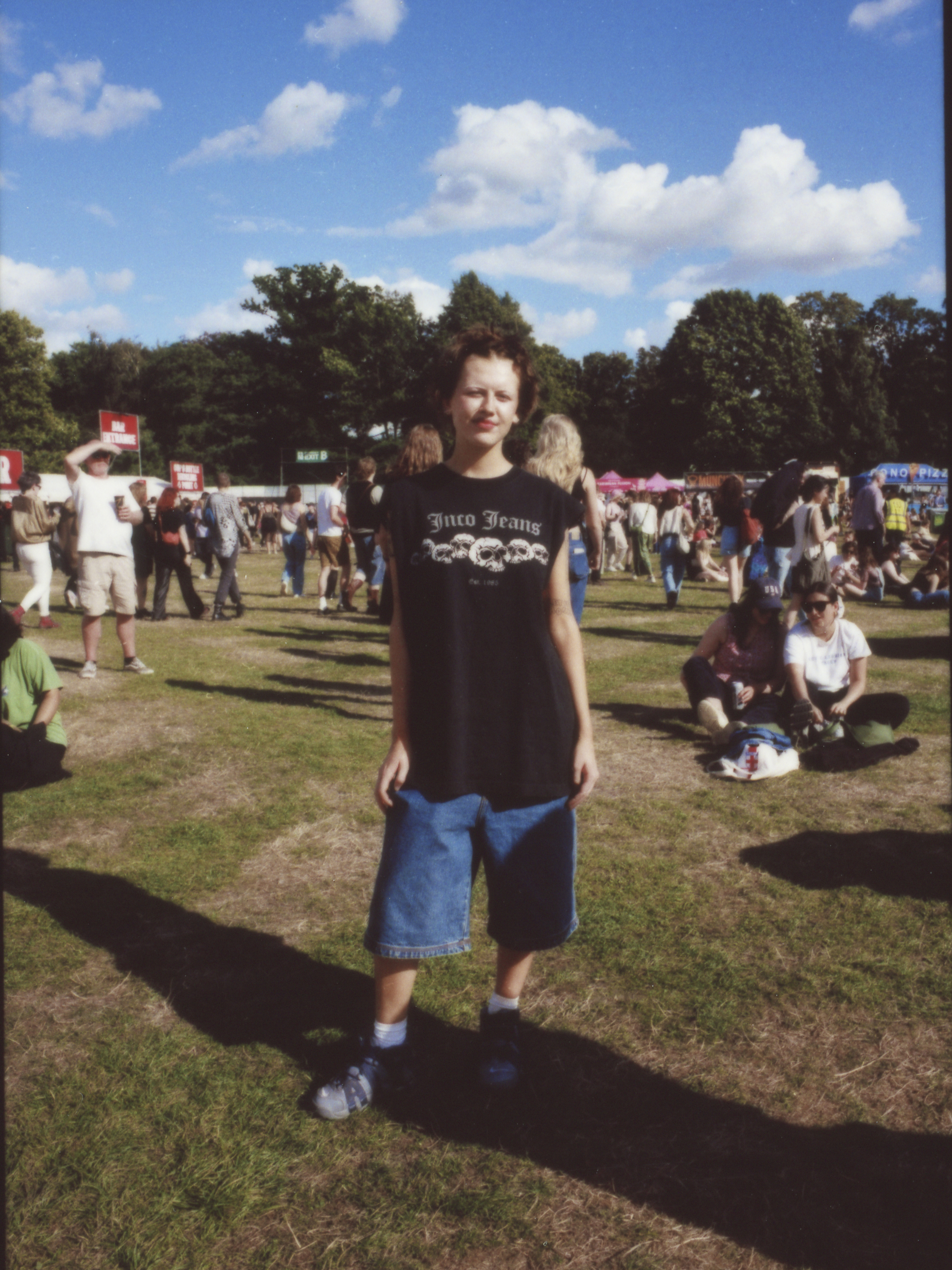 a young person wearing baggy denim shorts with an oversized tank top with skulls on it. they have short spiky hair and are squinting in the sun photographed by Jody Evans
