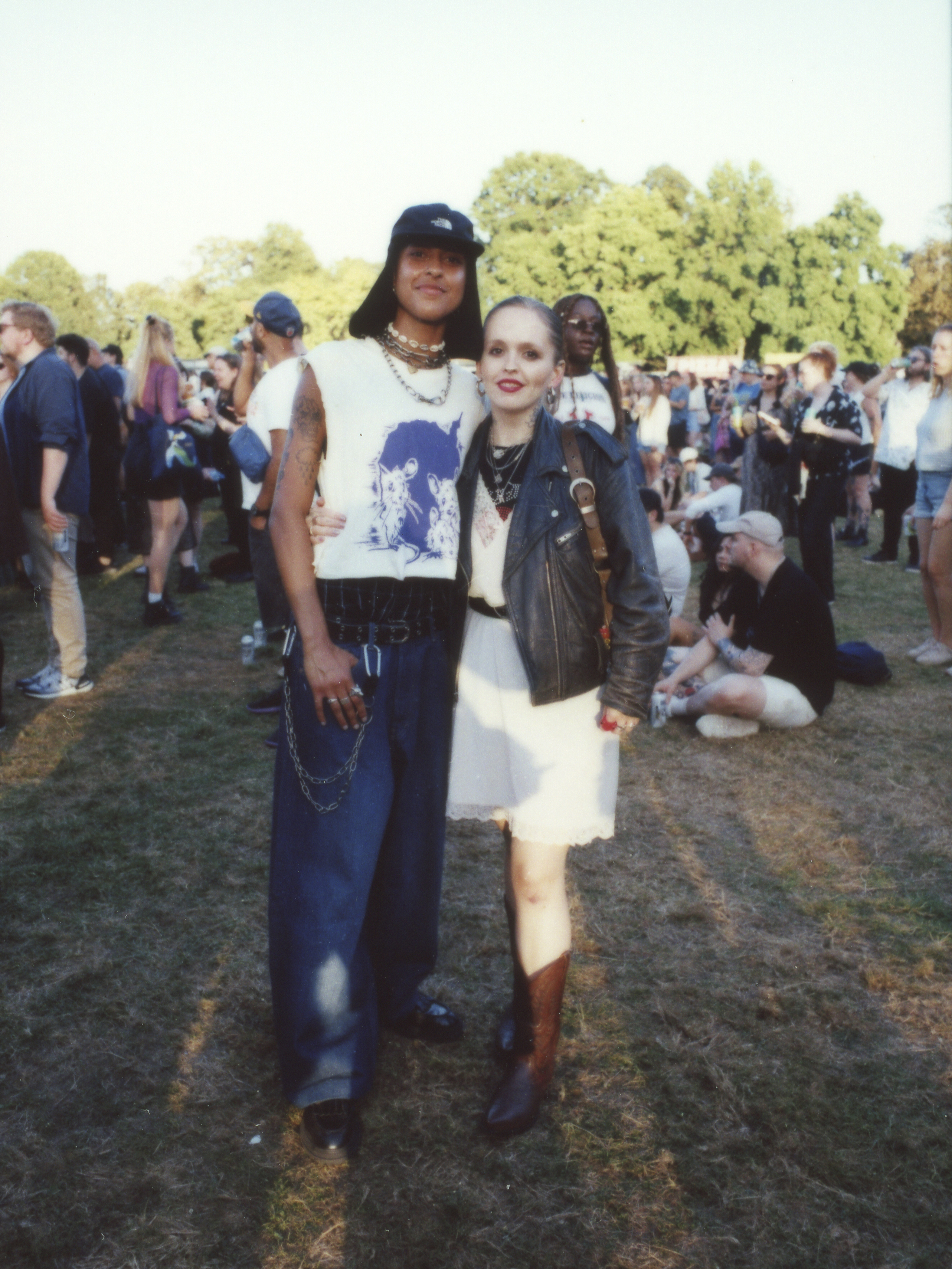 a couple with their arms around each other posing for a photo; one is wearing jeans with chains and a boygenius merch tee; the other a leather jacket over a dress photographed by Jody Evans