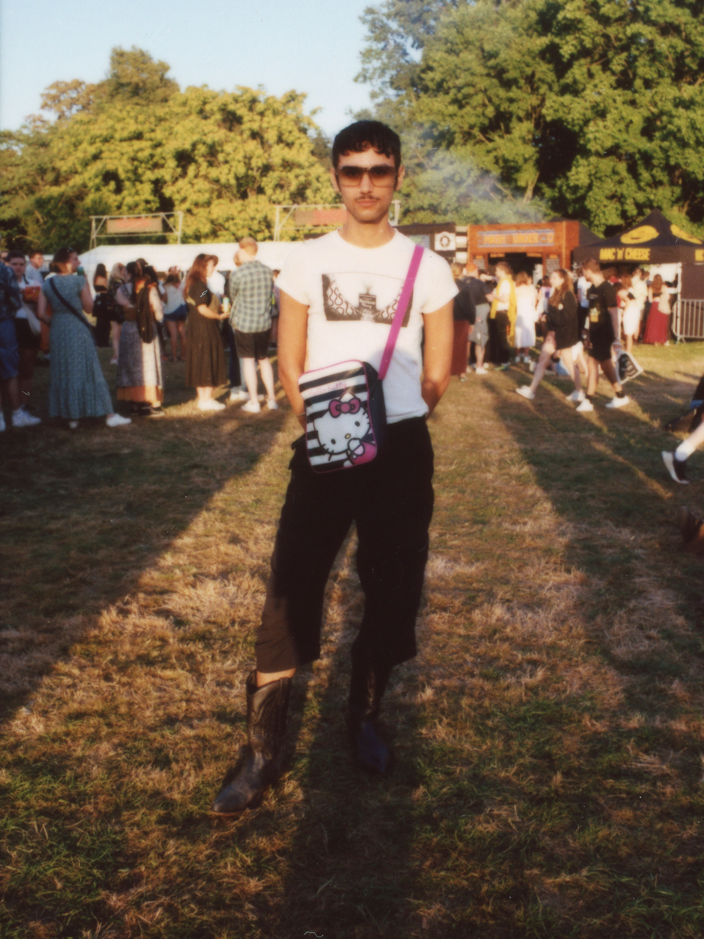 a festival-goert with facial hair wears a tshirt with cartoon breasts on it, a hello kitty messanger bag, jeans and cowboy boots photographed by Jody Evans