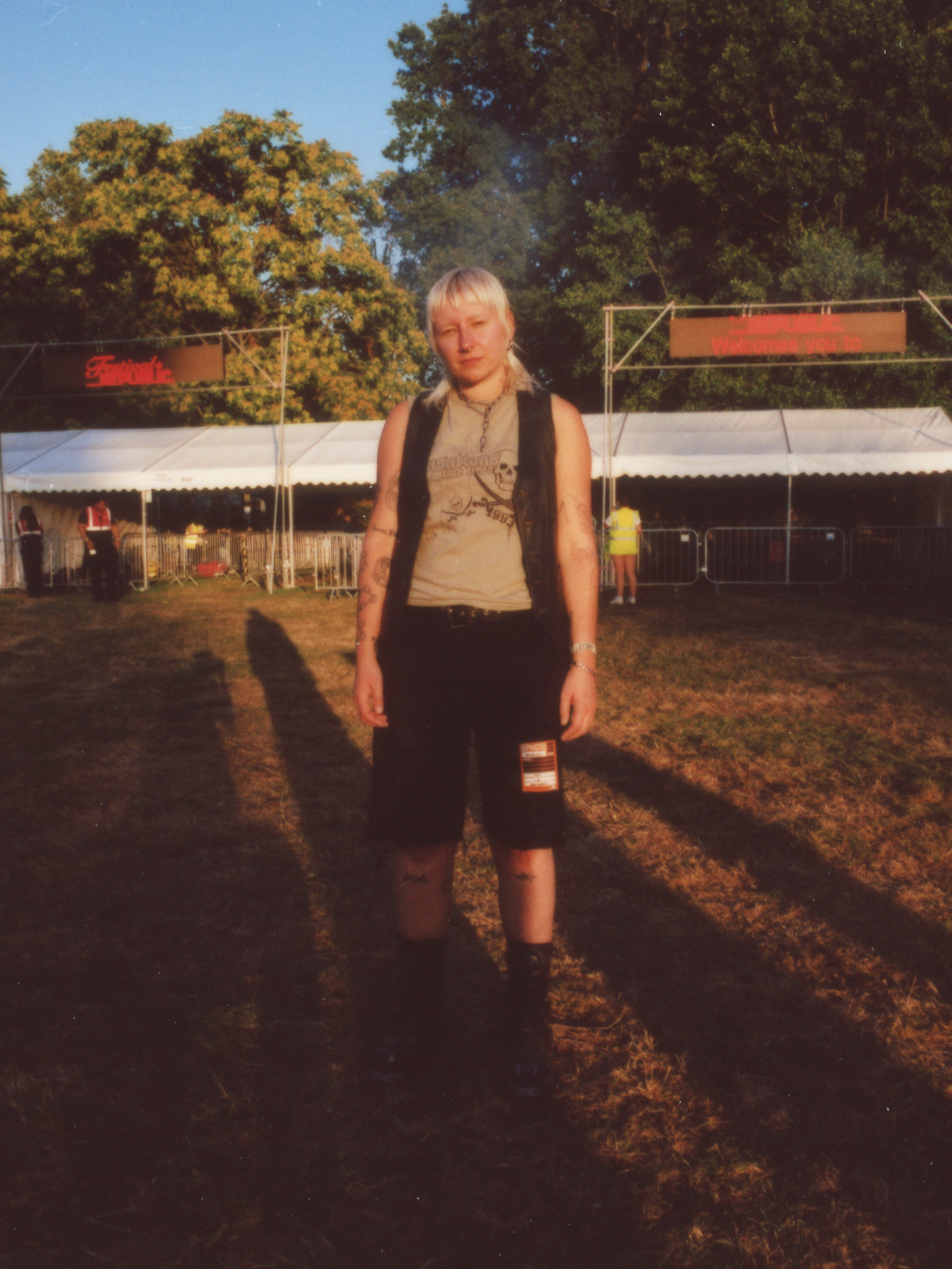 the photographer jody evans stands in golden hour light wearing jean shorts with a sticky on them; a skeleton tank top and an open leather vest; they have a bleach blonde mullet