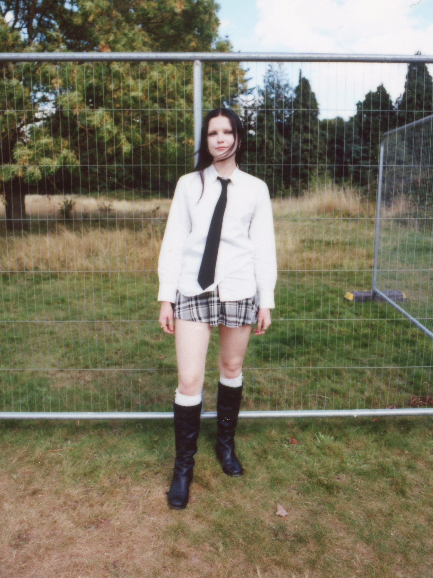 a festival-goer wearing knee-high boots with a short checked skirt, shirt and black tie stands in front of a metal gate photographed by Jody Evans
