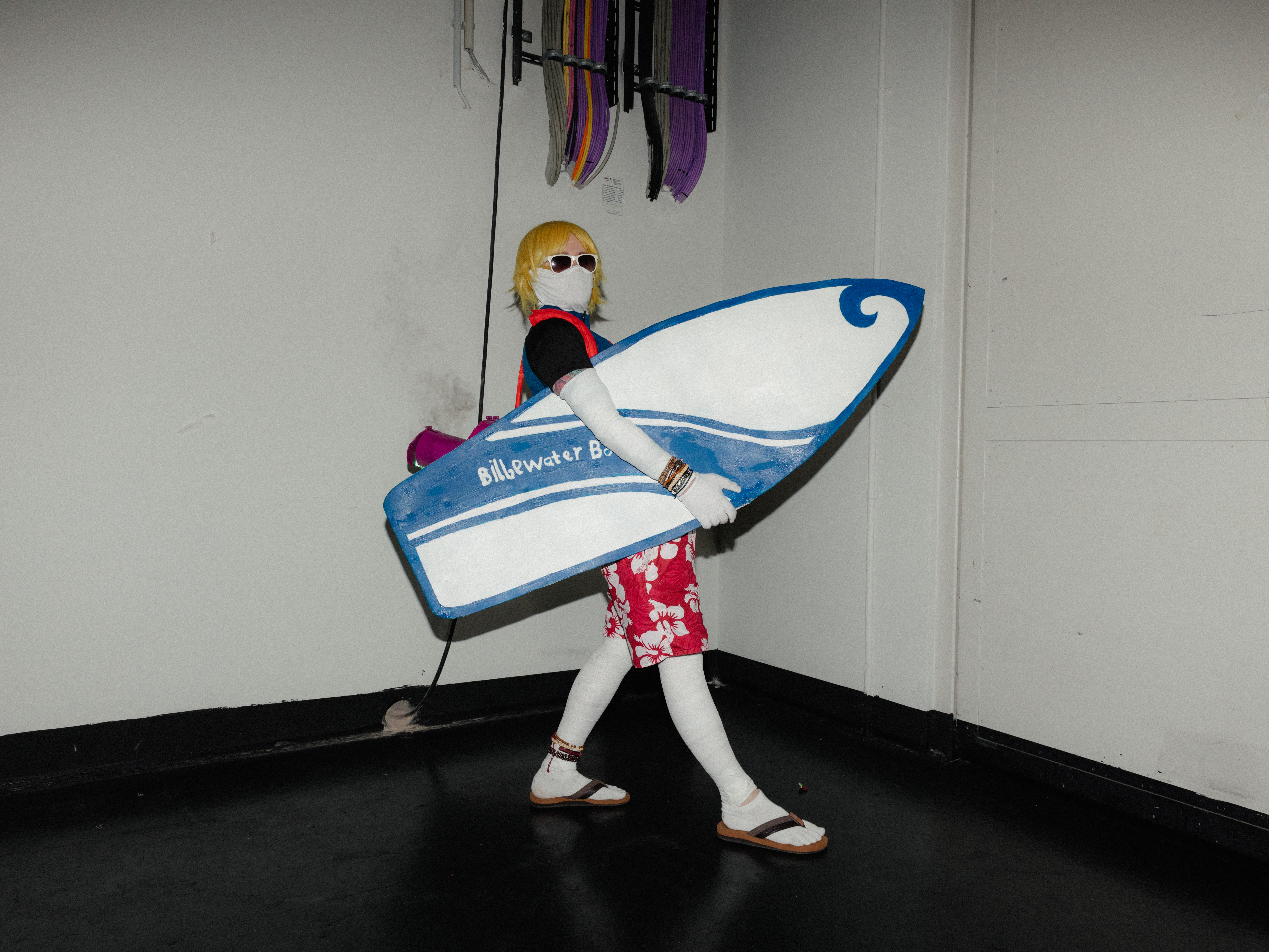 Ein Mann mit blonden Haaren, weißen Strumpfhosen, roter Boardshort hält ein blau-weißes Surfbrett