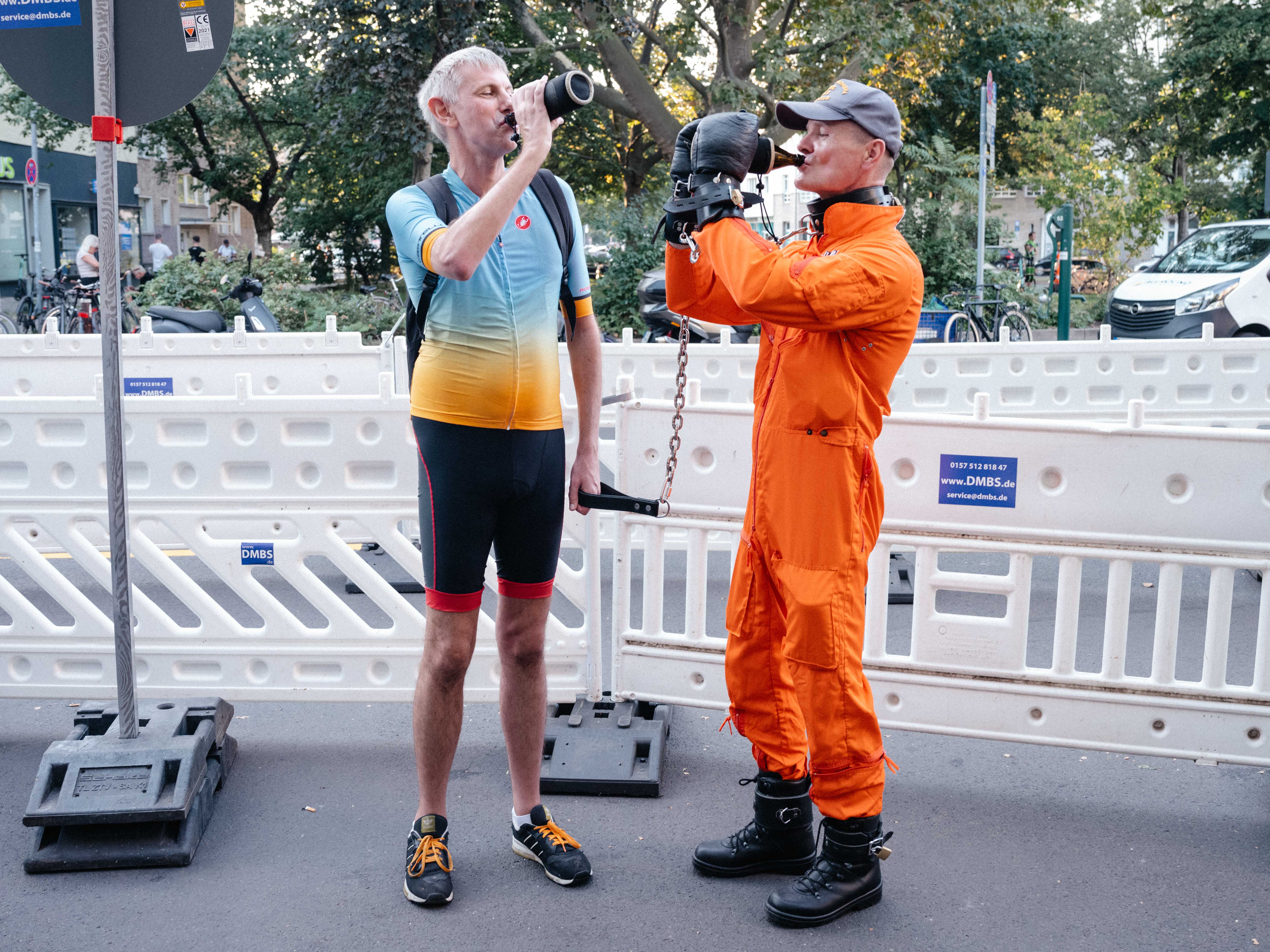 Marc trägt bei einem Fetisch-Event ein Rennradoutfit und führt an einer Leine einen Mann, der einen orangefarbenen Overall und Handfesseln trägt