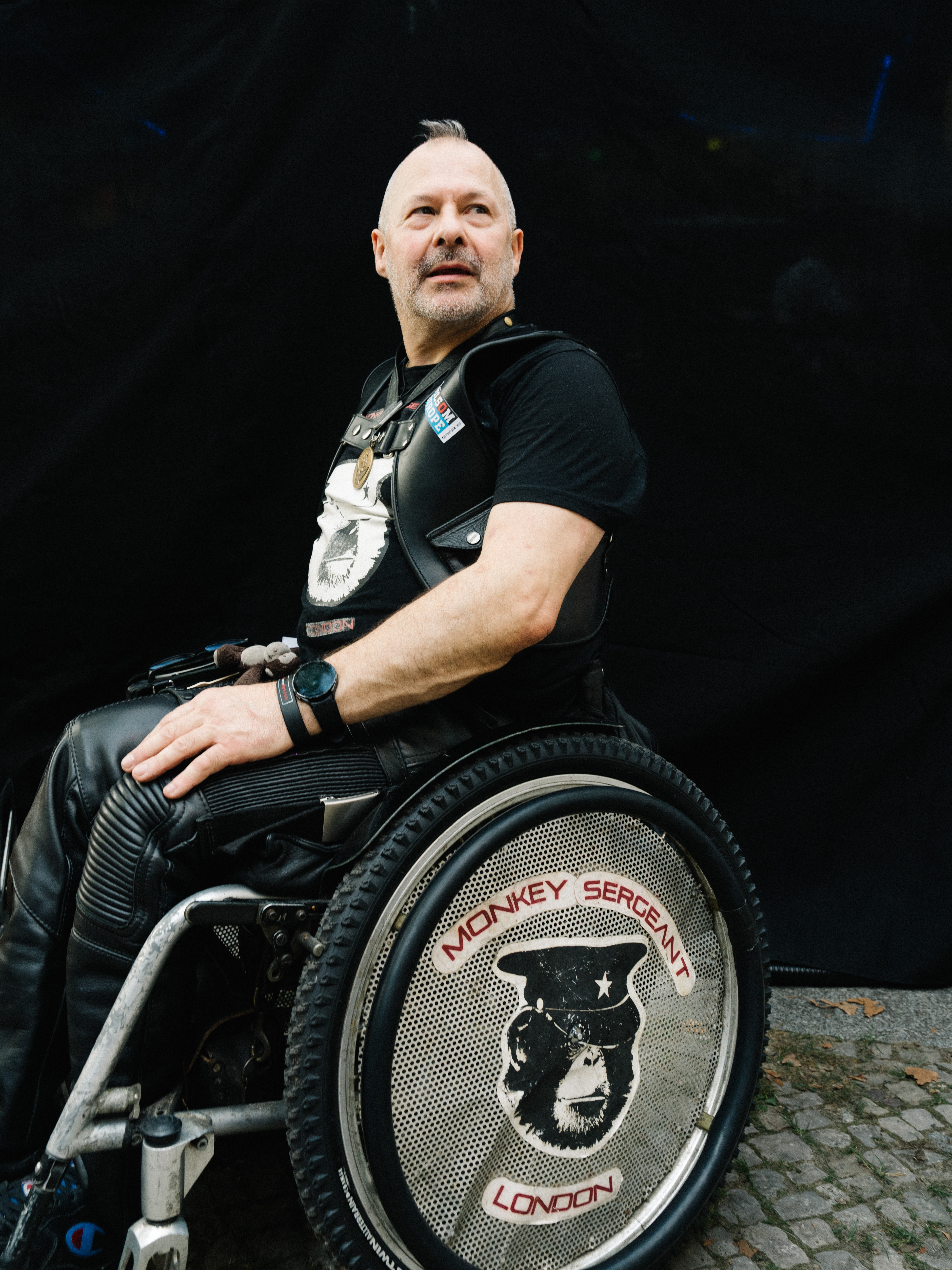 Ein Mann trägt schwere Lederkleidung, er sitzt in einem Rollstuhl auf dessem Rad steht: 
