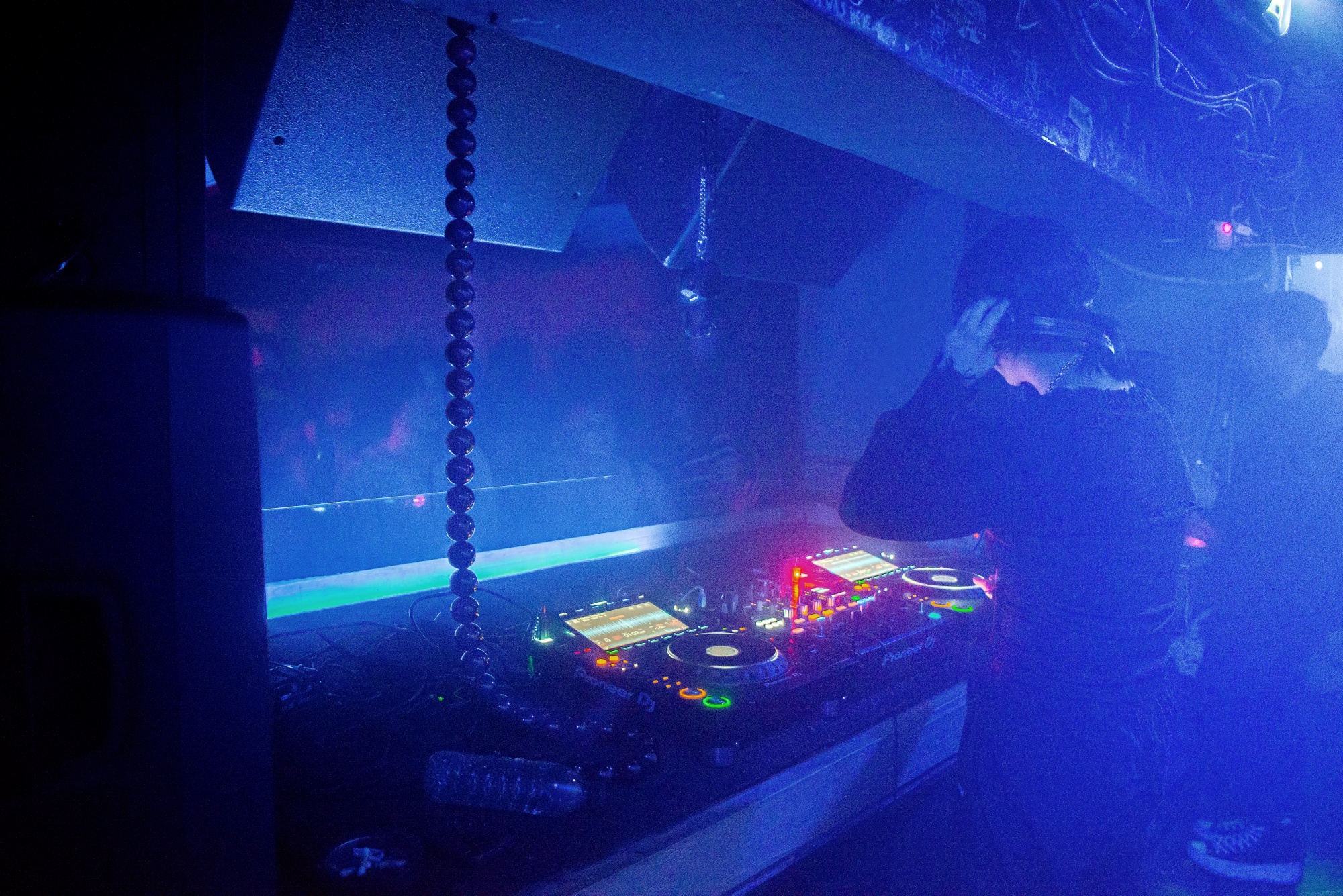 Ein DJ steht in blauem Licht in einer DJ-Booth vor CDJs und einem Mixer