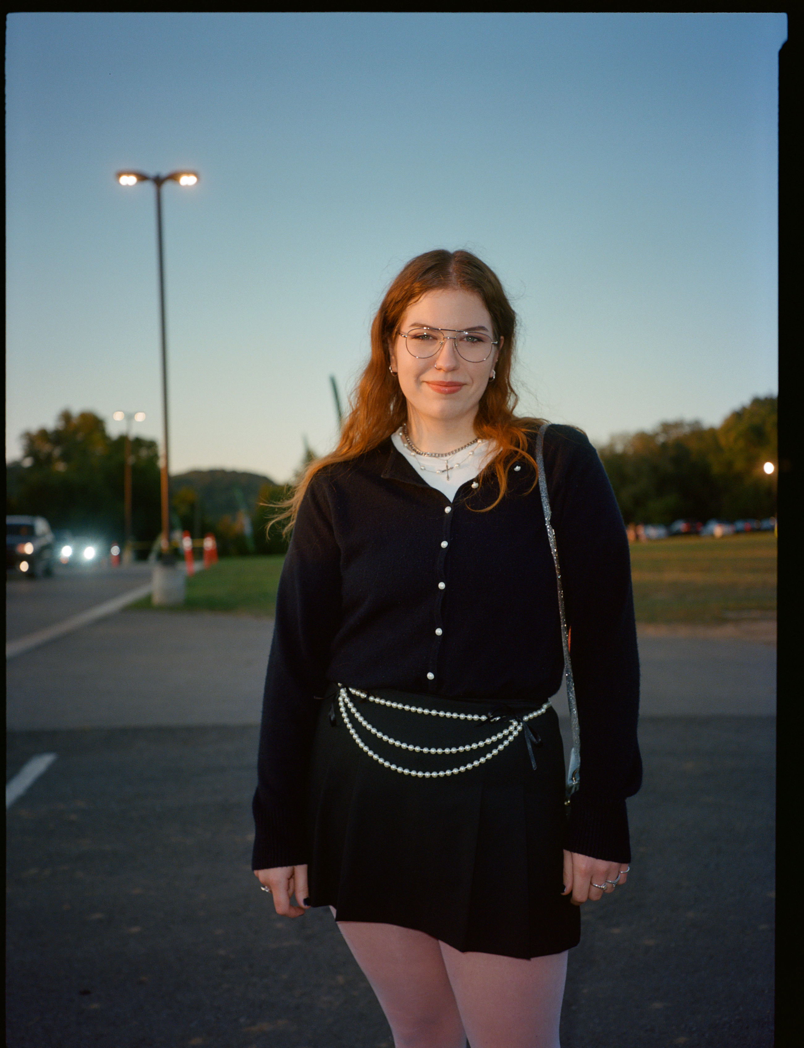 a lana del rey stan stands in a car part. she has long auburn hair and framed glasses. she wears a black cardigan and black skirt with a belt made of pearls
