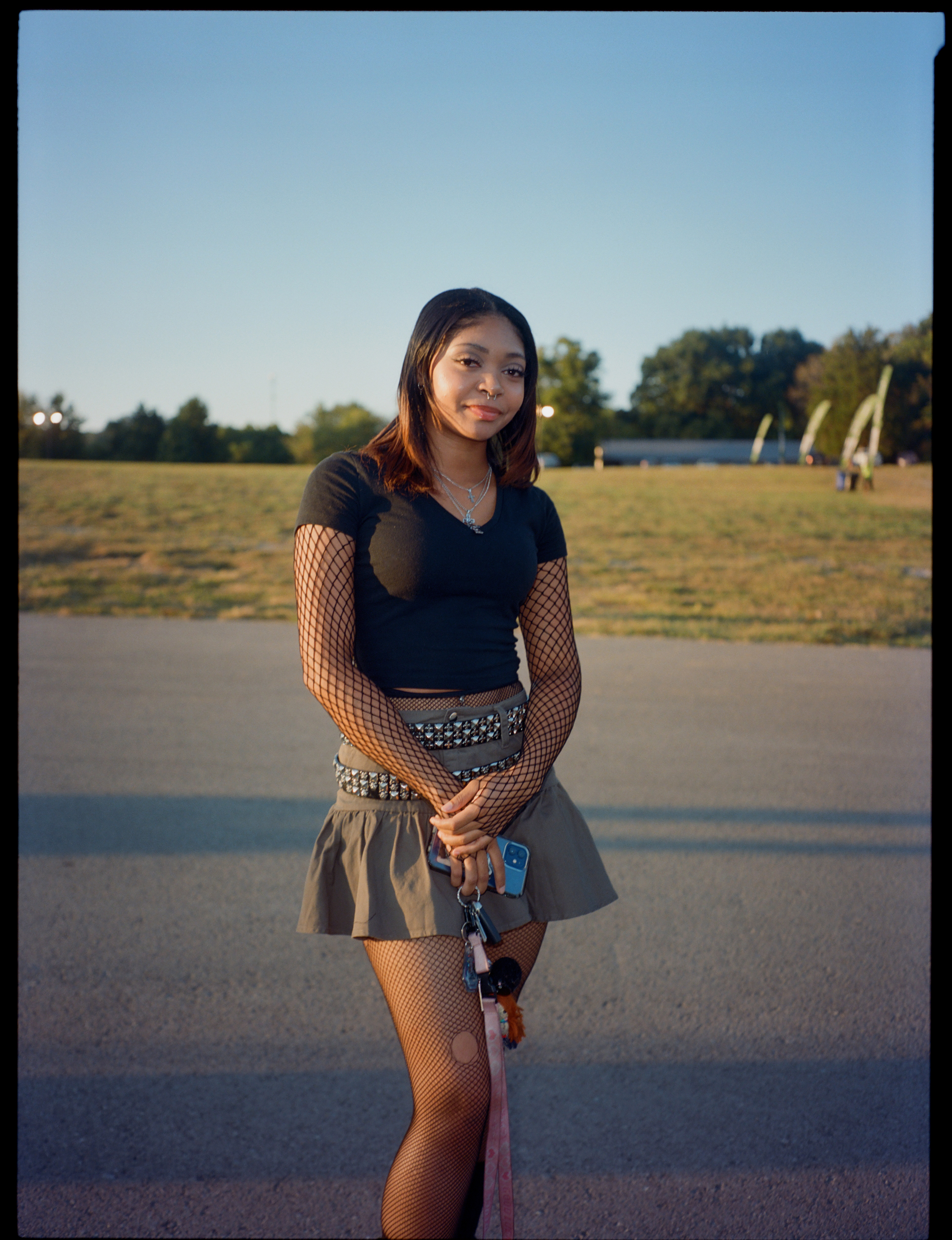 a lana del rey fan in tennessee wears a black t-shirt with fishnet sleeves, and a khaki peplum skirt with studded belt. she has shoulder length hair and is smiling
