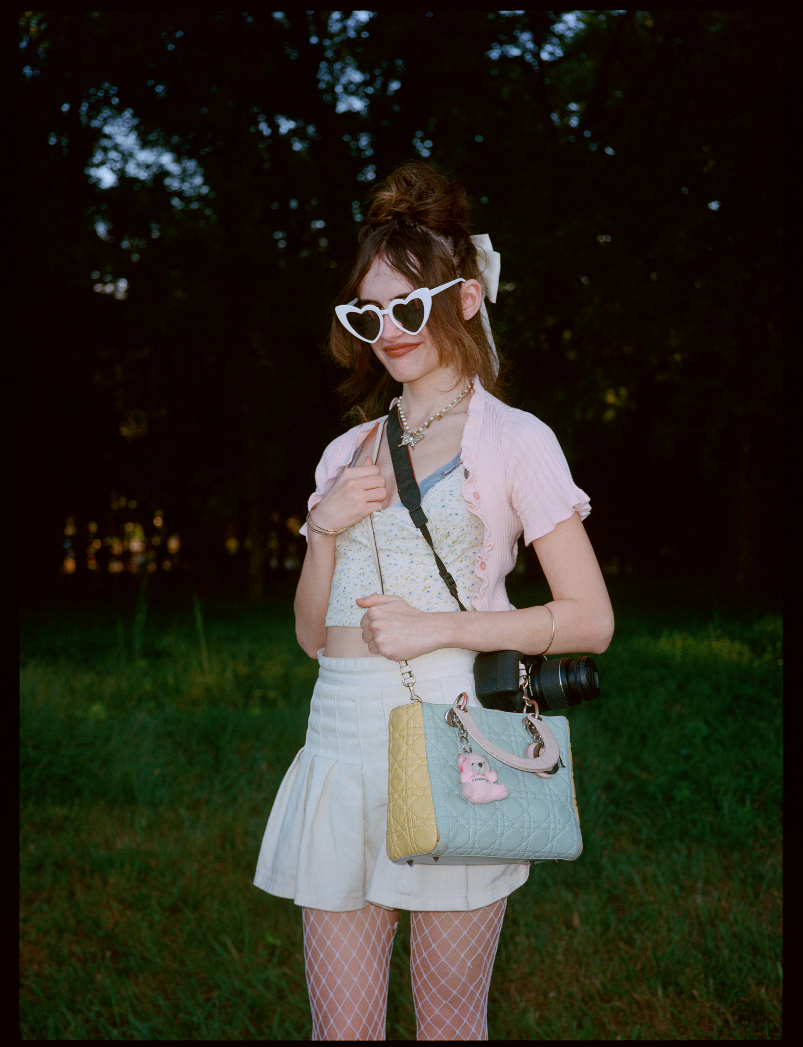 a lana del rey fan wears a cropped blazer and cardigan and a white peplum skirt. she carries a pastel-coloured handbag and wears glasses shaped like love hearts