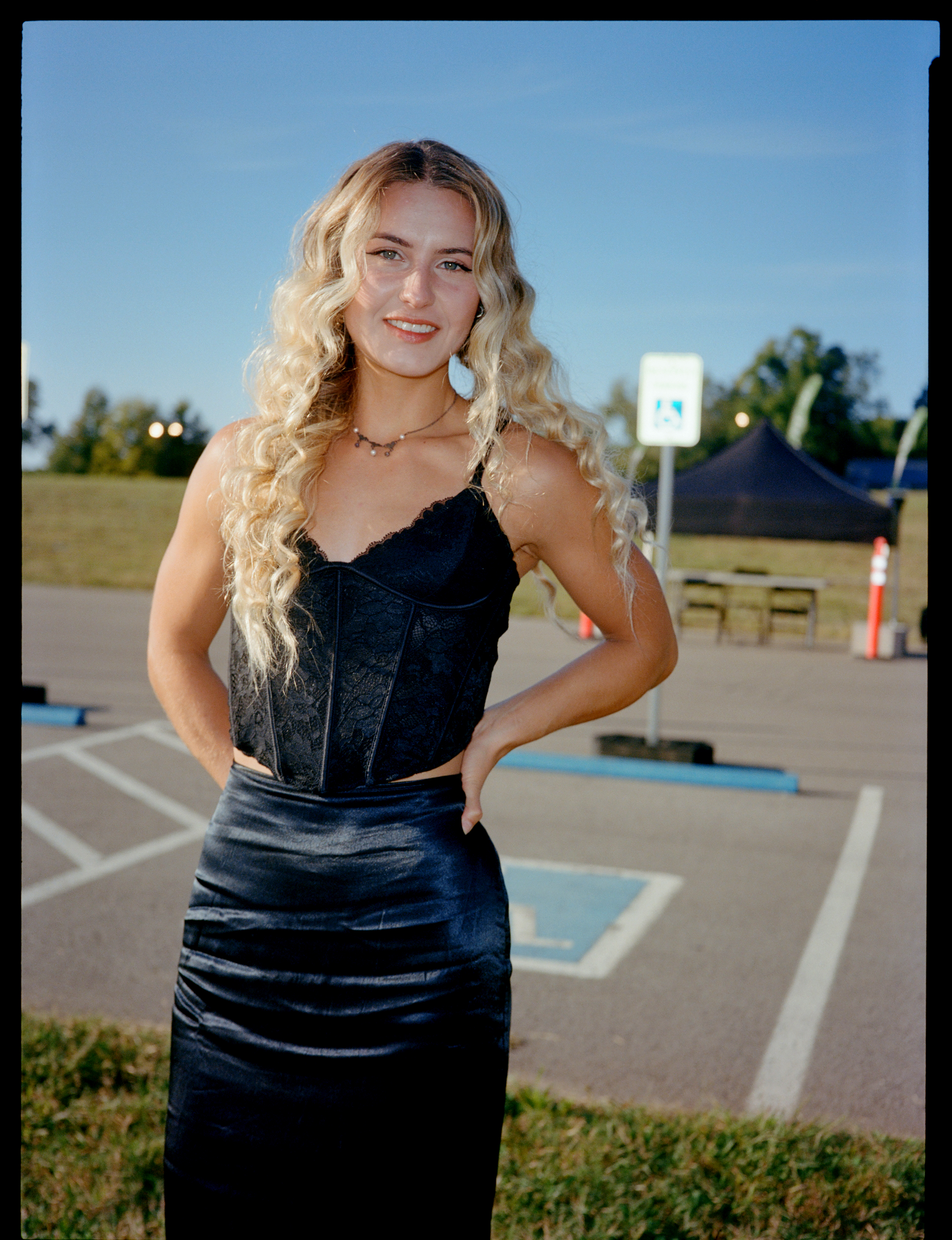 a lana del rey fan stands in a car park wearing a black corset lace top and a black skirt. she has blonde hair and is smiling. 