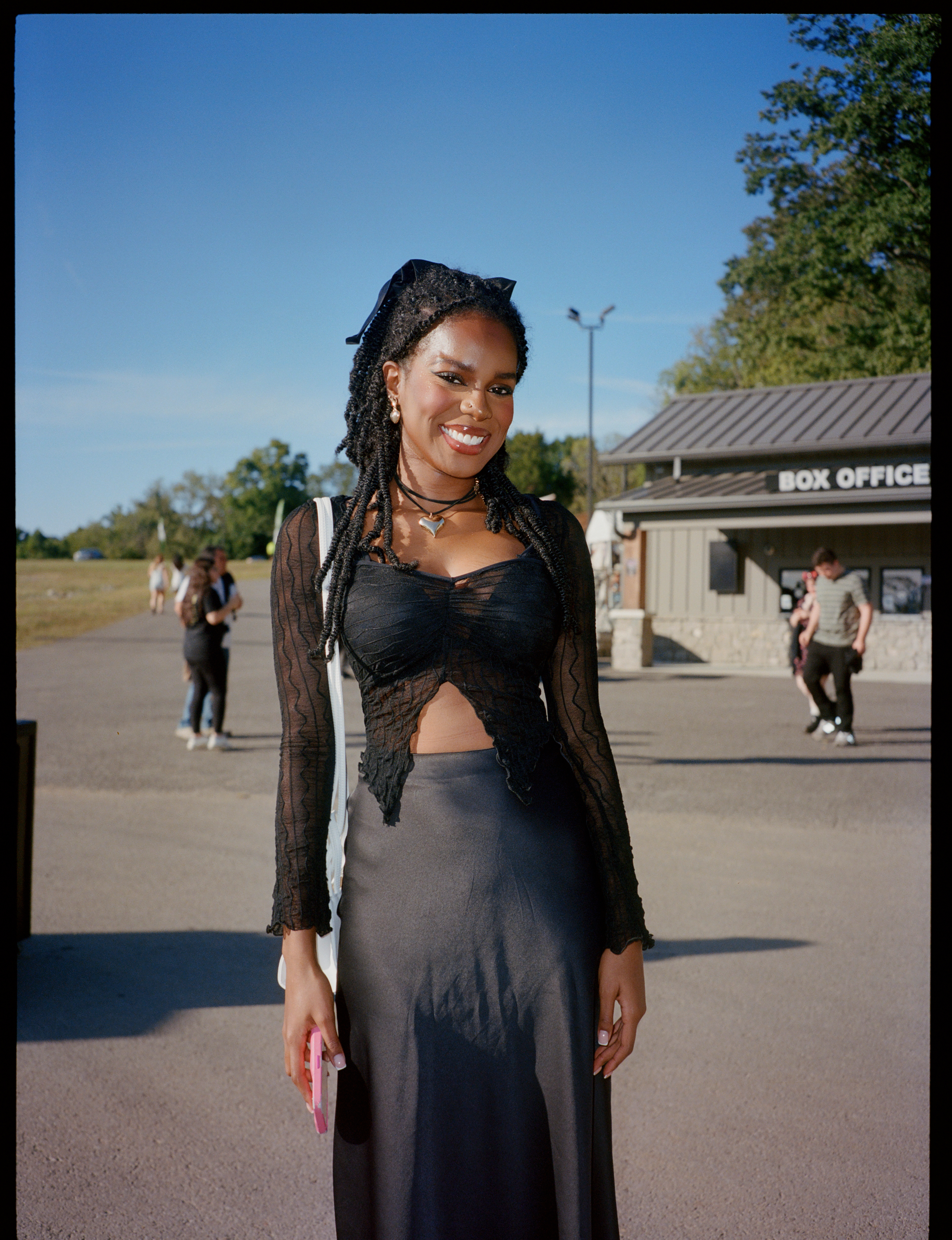 a lana del rey fan stands outside the pavilion at one of her shows. she has black braids in her hair and is wearing a black skirt and a sheer black top.