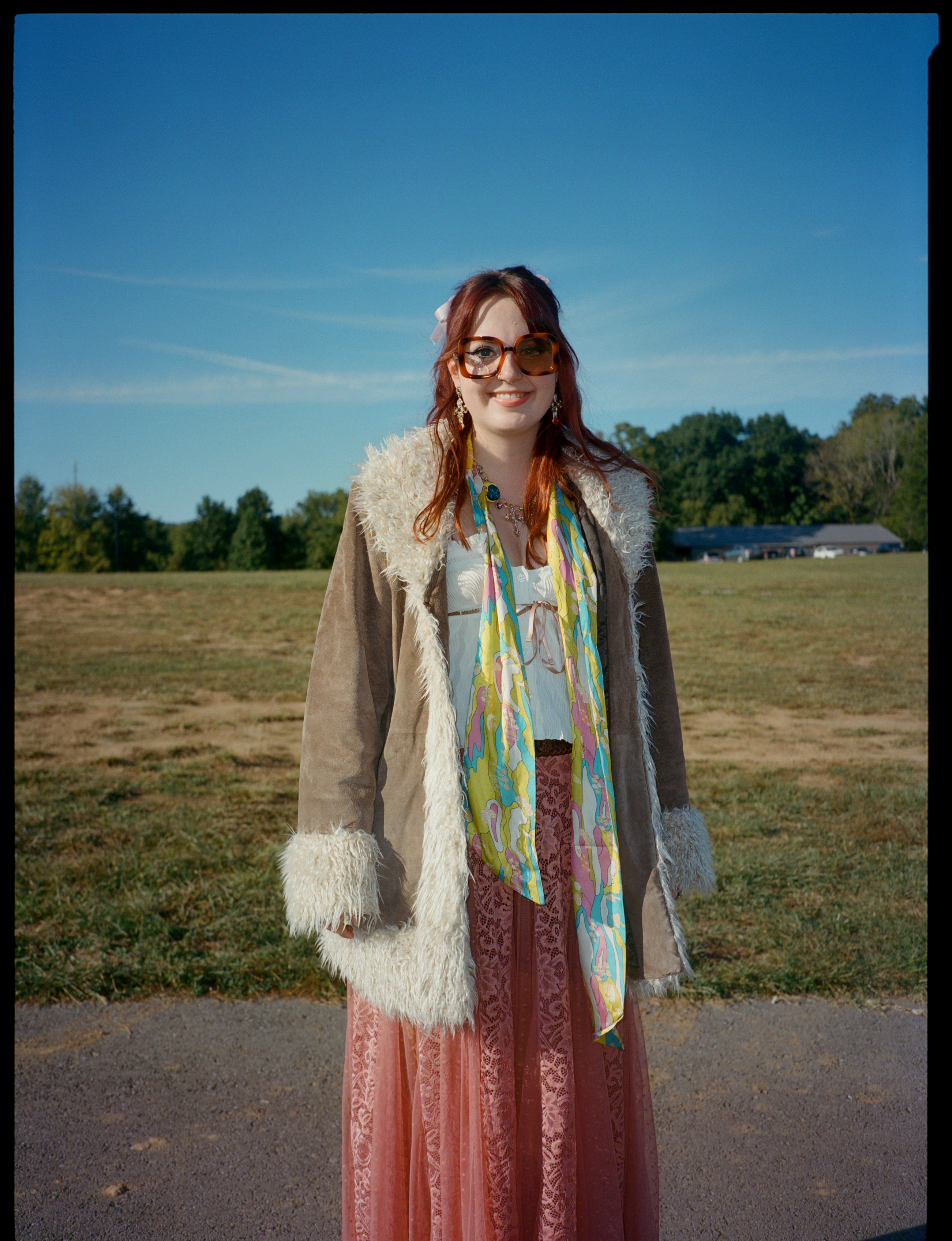 a lana del rey fan wears a fur-lined jacket and wide rimmed sunglasses. she has a long pink skirt on and has a multicolour scarf round her neck.