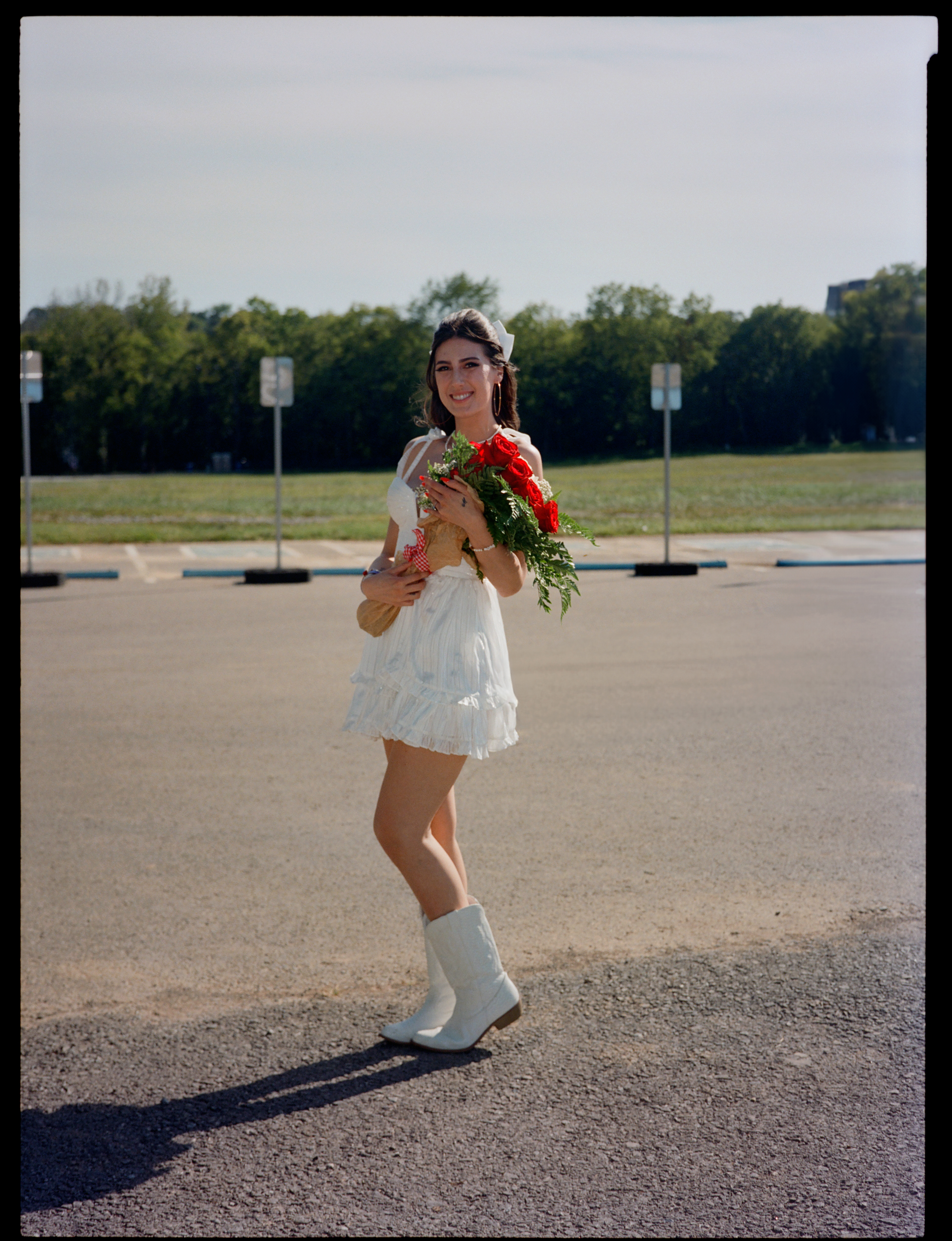 a lana del rey fan stands in a car park wearing cow boy boots, a white dress, and carrying a bouquet of red flowers 