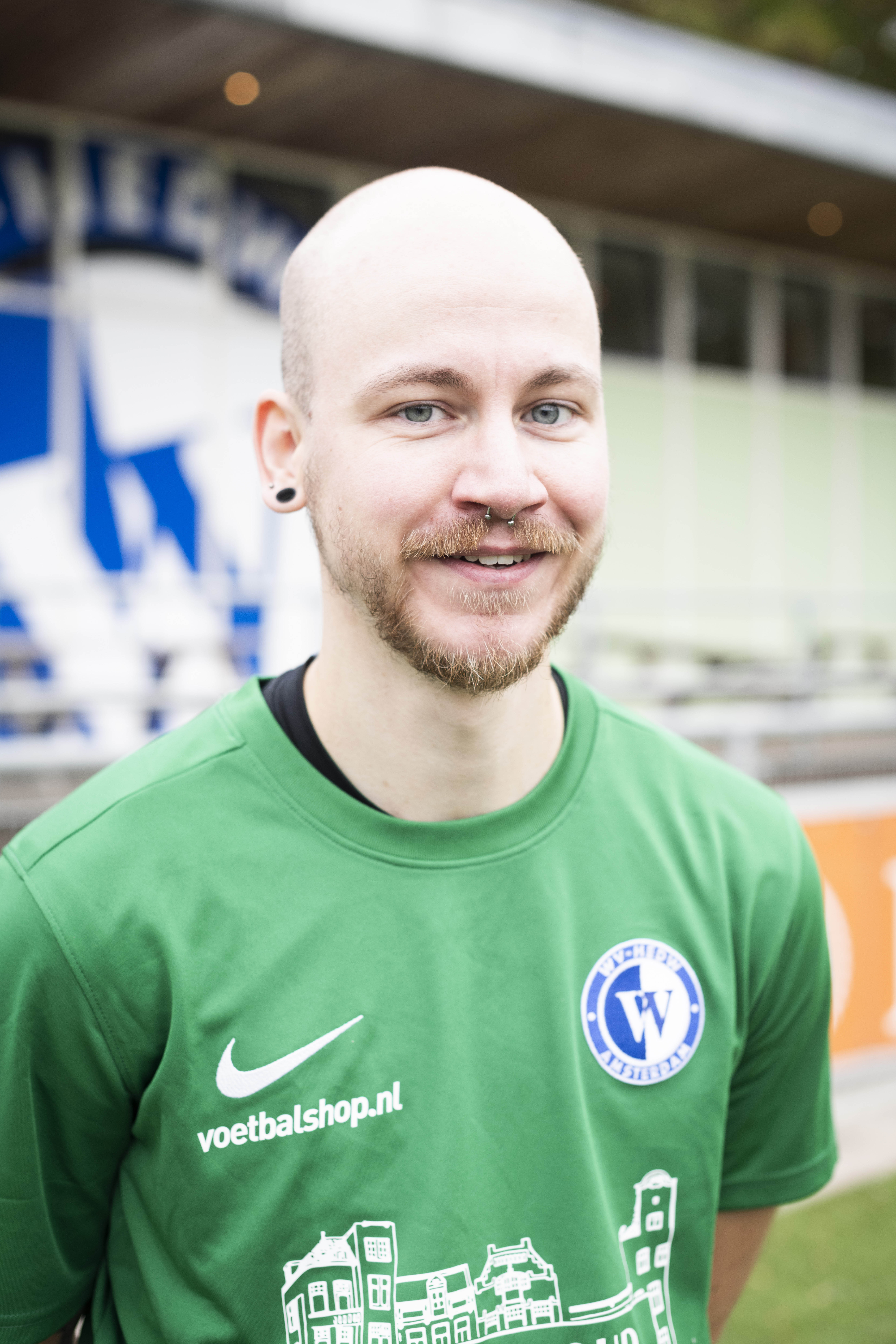 Portrait of a football player wearing a green football jersey on the pitch, he has piercings in his nose and ear, looks smiling at the camera