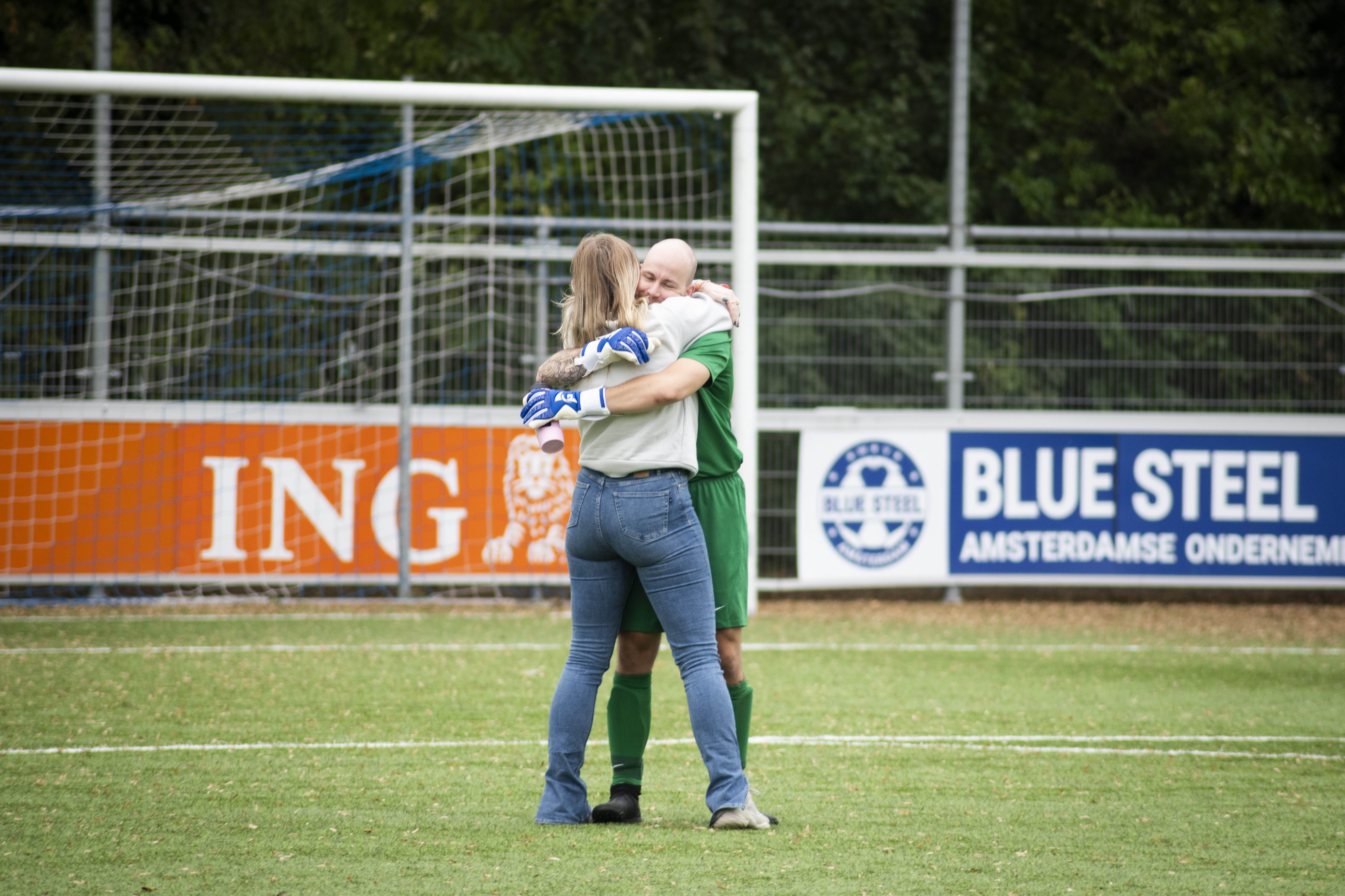 two people hugging on a football field. One wears a green football jersey and the other wear blue jeans and a white jumper. There's an ING blank and Blue steel ad in the background