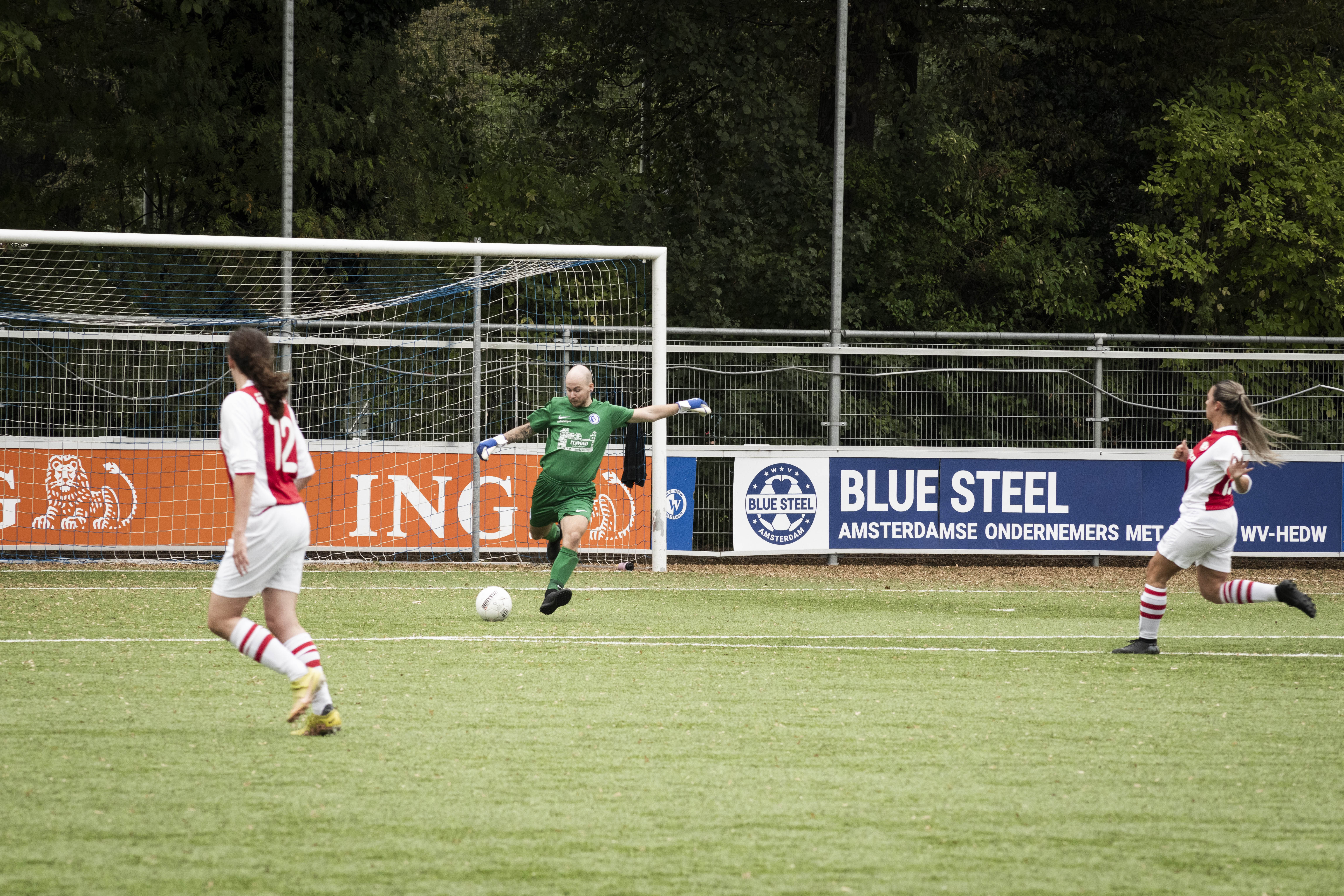 action photo of three people playing football, the keeper wears a green jersey and is about to kick the ball and two opponents are running in a red and white jersey