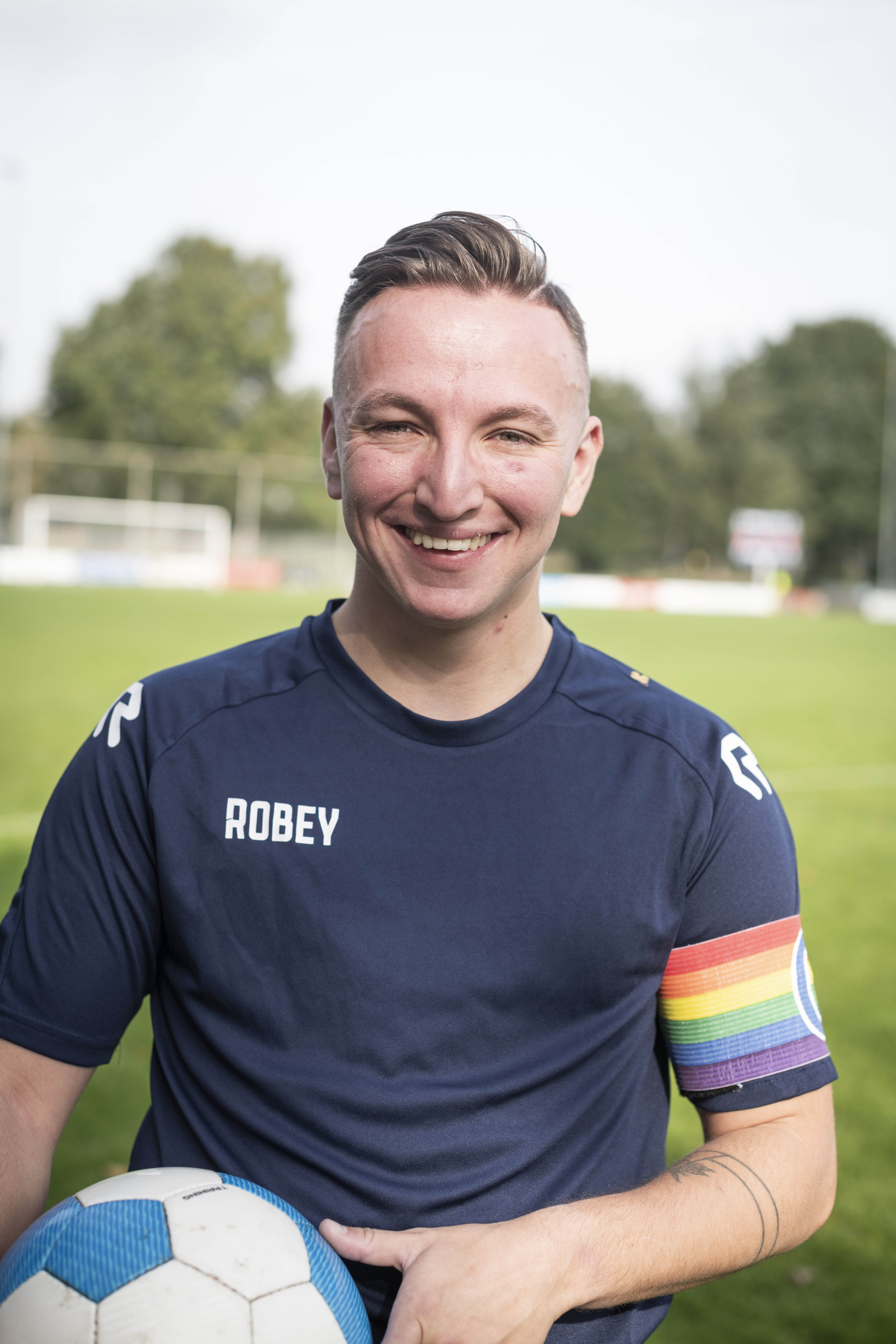 portrait of a football player standing on the pitch. He's wearing a blue shirt with Robey written on it, a rainbow armband and they're carrying a white and blue ball