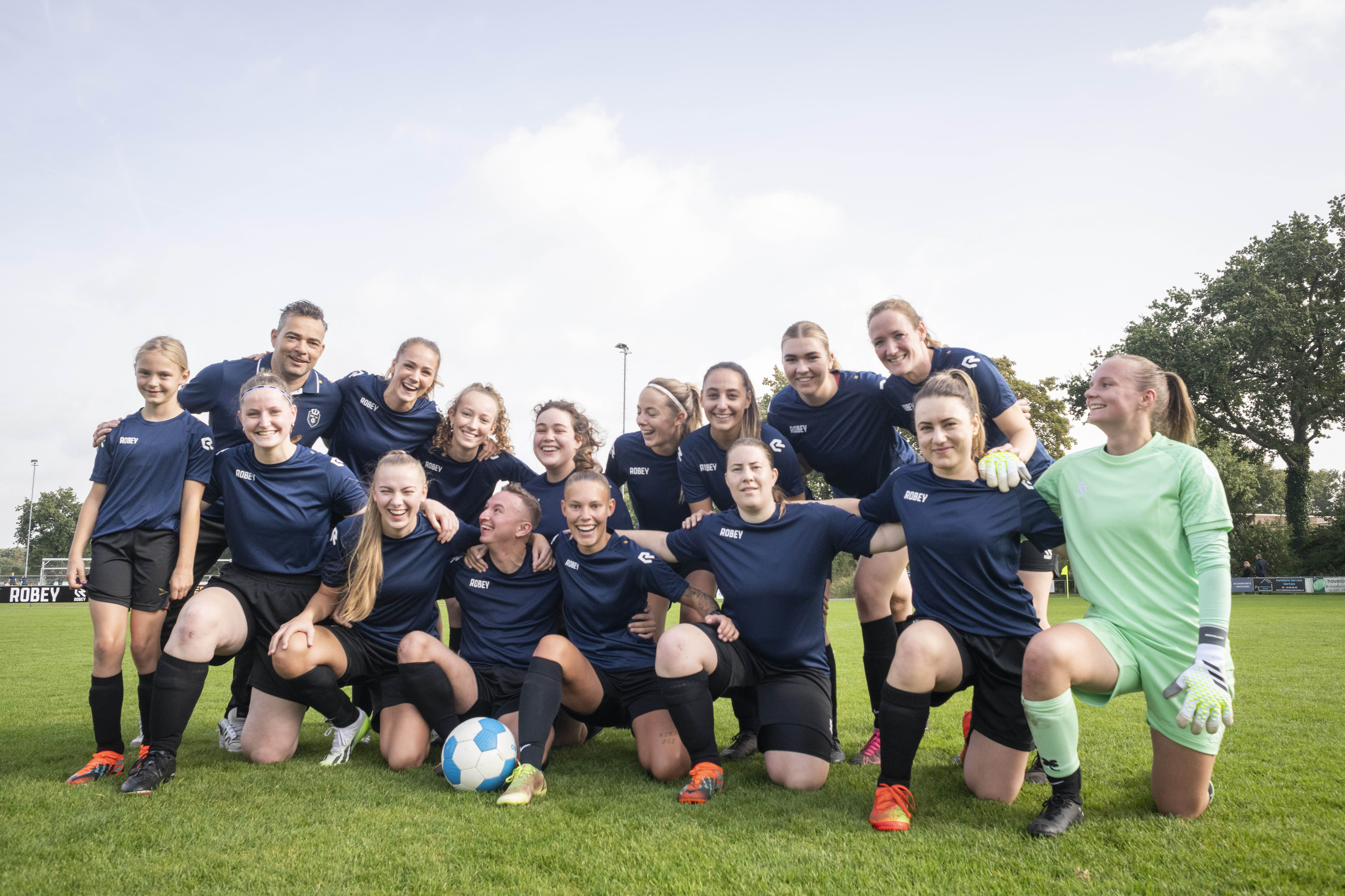 group photo of a women and queer football team on the pitch, they're wearing black and dark blue football jerseys except for the goal keeper who's dressed in green 