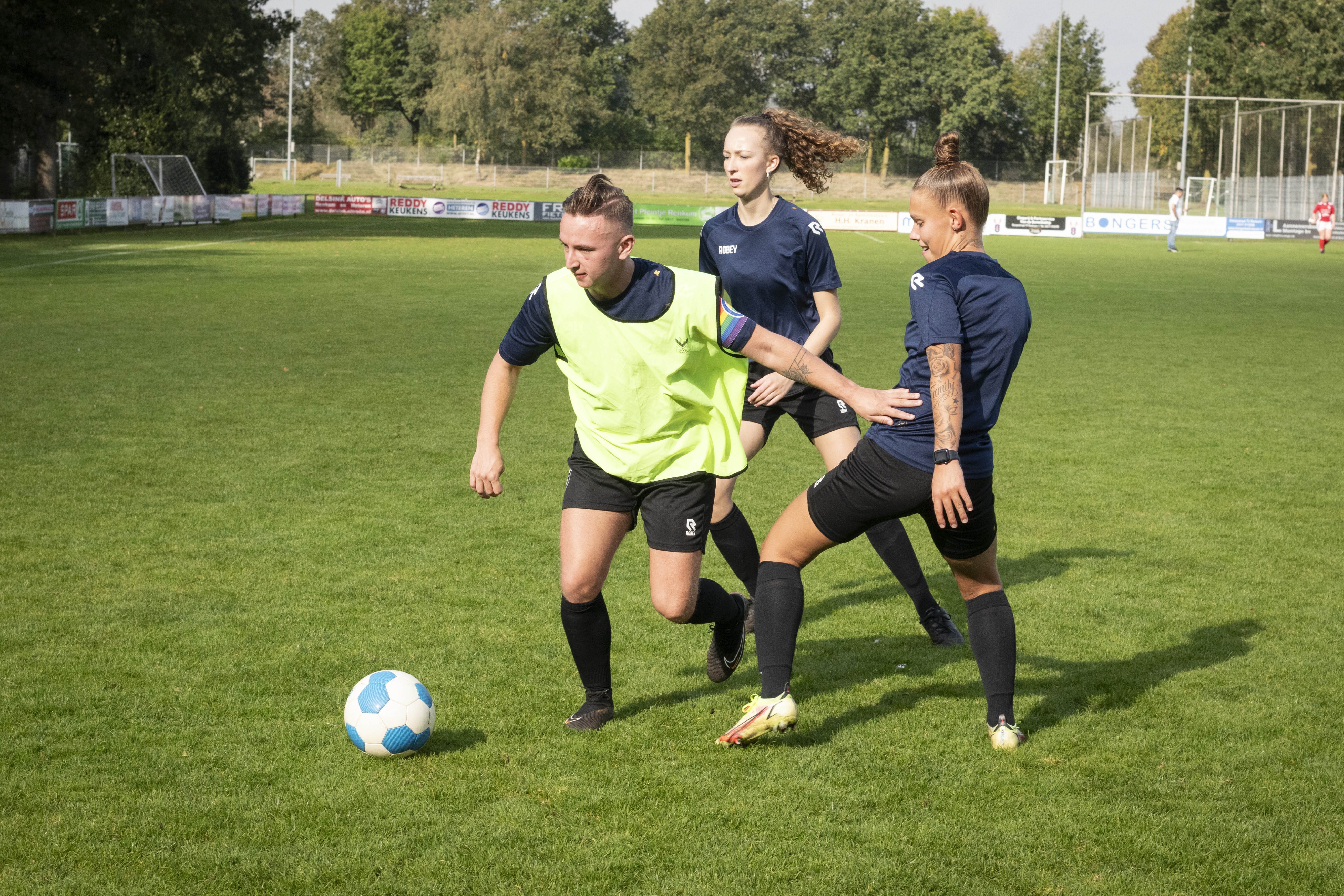 action shot of three people playing football on a sunny day, they're wearing a black and dark blue jersey and one of them has a yellow jersey on top